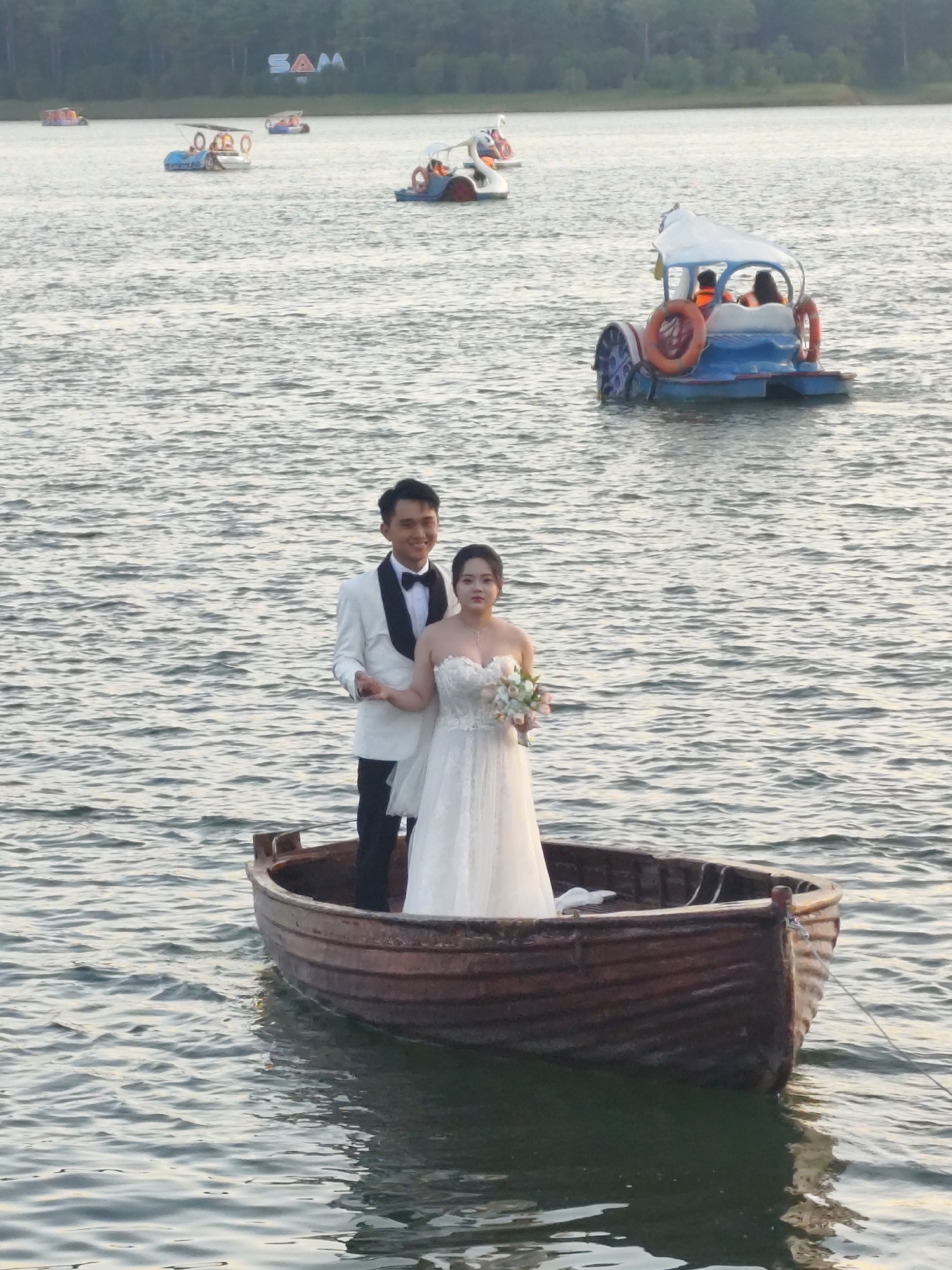 An outdoor, medium shot captures a wedding couple posing for photos in a small, wooden rowboat on a body of water. The couple is positioned towards the right side of the frame, with the woman to the right of the man. The man, dressed in a white tuxedo with a black lapel and a black bow tie, smiles directly at the camera. The woman, wearing a strapless white wedding gown and holding a bouquet of light-colored flowers, looks slightly off-camera with a neutral expression. The boat is brown and has visible wood grain. The water is rippled, reflecting the sky and surrounding environment. In the background, several other pedal boats are visible on the water, and a tree-lined shore stretches across the horizon. The sky is overcast and hazy.