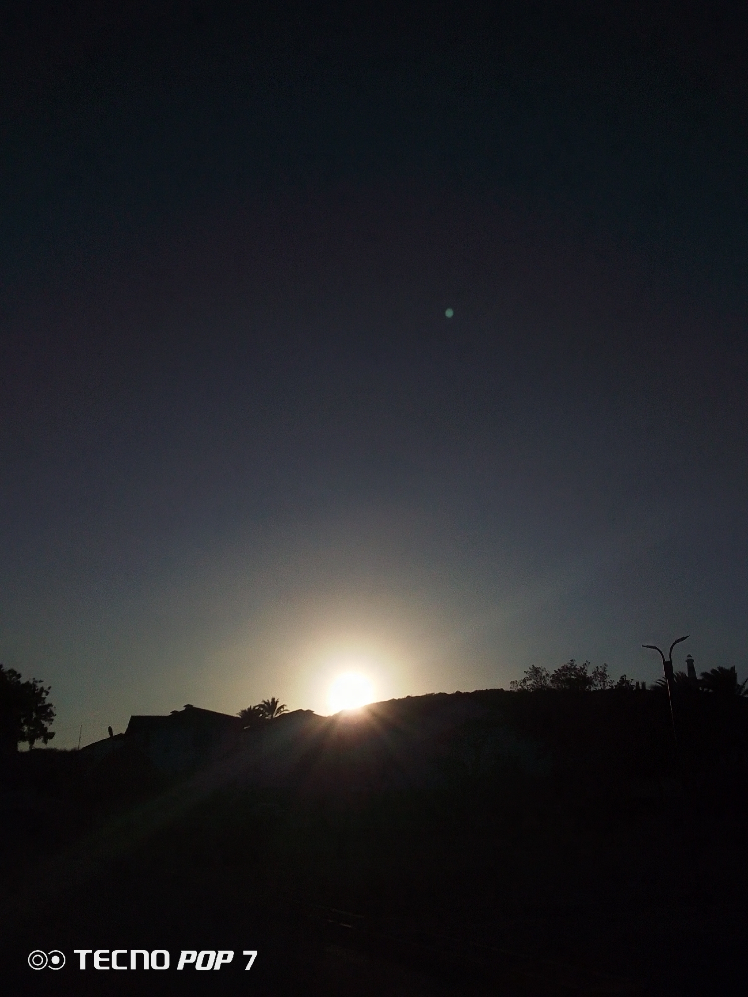 The image shows a sunset in Pedro González, Venezuela. The sun is positioned on the horizon, casting light above a dark hill and vegetation. The sky is dark, with a single light spot in the upper part of the image. A building is on the hill, and a light pole is visible on the right. The camera is marked as "TECNO POP 7".