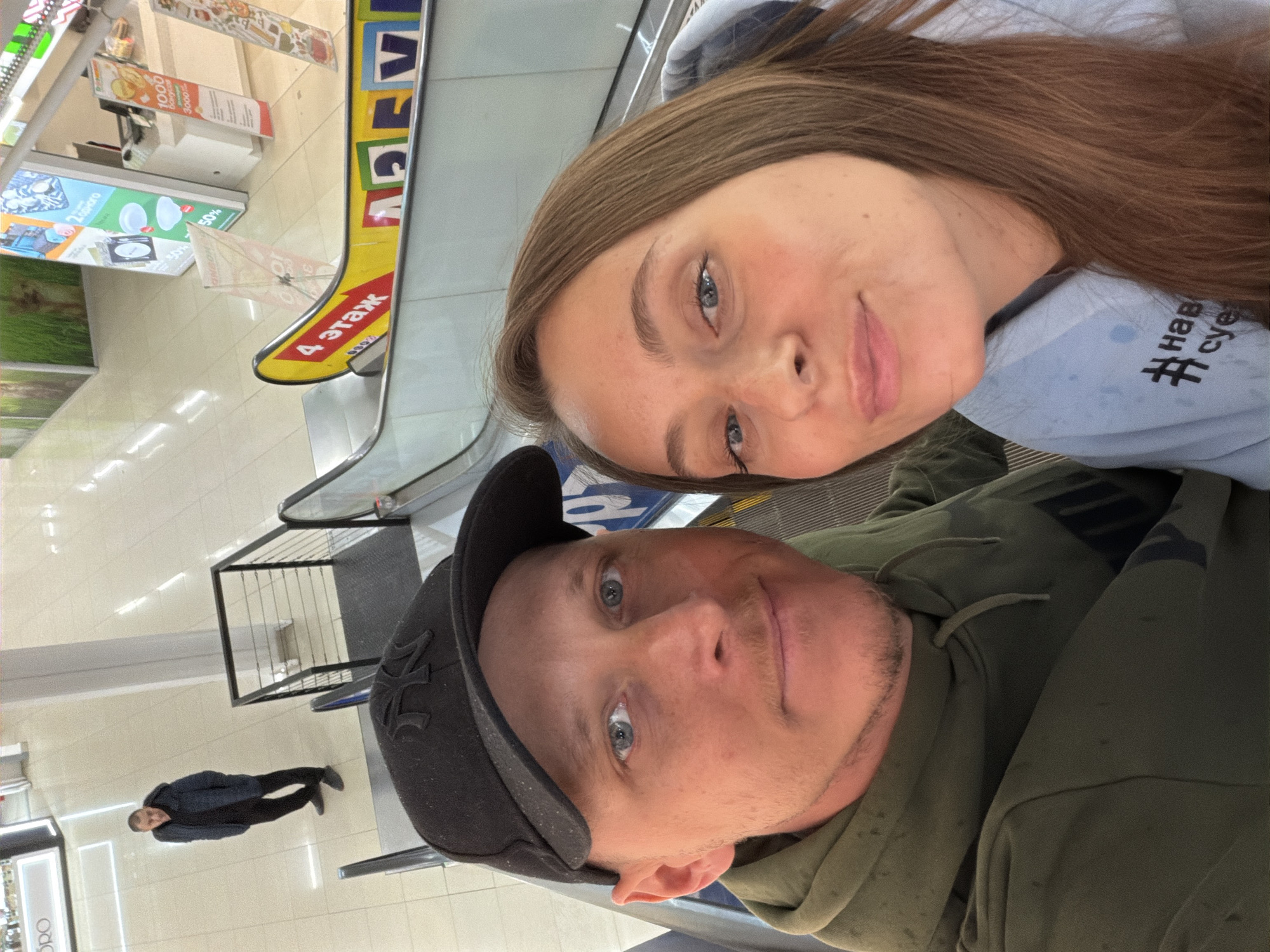 The image depicts a selfie taken by a man and a woman on an escalator in a multi-level shopping center in Birobidzhan, Russia. The man, positioned on the left, has blue eyes, fair skin, and light stubble, wearing a black baseball cap with a white "NY" logo and an olive green hooded sweatshirt. The woman, to his right, has long brown hair, blue eyes, and fair skin, wearing a light blue hooded sweatshirt with black Cyrillic text partially visible, including "# нав суе". Both individuals are looking directly at the camera.
In the background, on the floor below the escalator, a third male individual, dressed in a dark blue or black jacket and dark pants, stands facing upwards towards the camera. The retail environment is brightly lit with white tiled flooring. A yellow sign affixed to the escalator's side railing indicates "4 этаж" (4th floor) in red Cyrillic script. Various promotional retail signs are visible in the background, displaying percentages (e.g., 30%, 50%), monetary values (e.g., 1000, 3000), and product imagery, with some partial Cyrillic text on them. A partial "ORO" sign is also discernible in the far background.