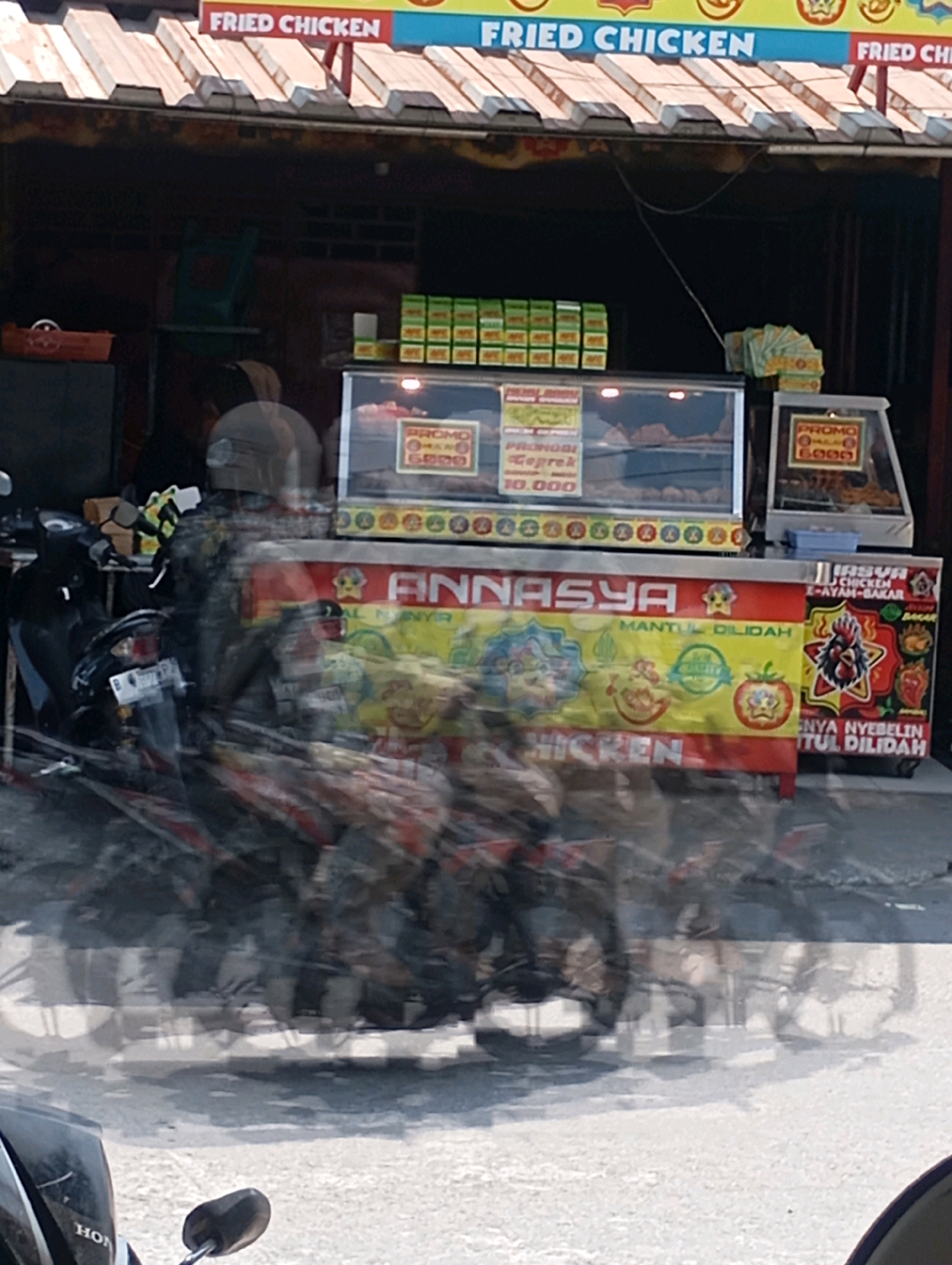 A fried chicken stall is captured in an outdoor scene, likely on a street or marketplace in Babelan, Indonesia. The stall features a display case with various food items, including fried chicken and what appear to be promotional boxes of food. Signs indicate "Fried Chicken" and "Promo" with prices like "6000". The stall is decorated with colorful banners, one prominently displaying the name "ANNASYA" along with images of fried chicken and vegetables. To the left of the stall, a motorcycle is parked, and a person wearing a helmet is partially visible, their form blurred due to motion. The ground appears to be paved, and the overall lighting suggests it is daytime.