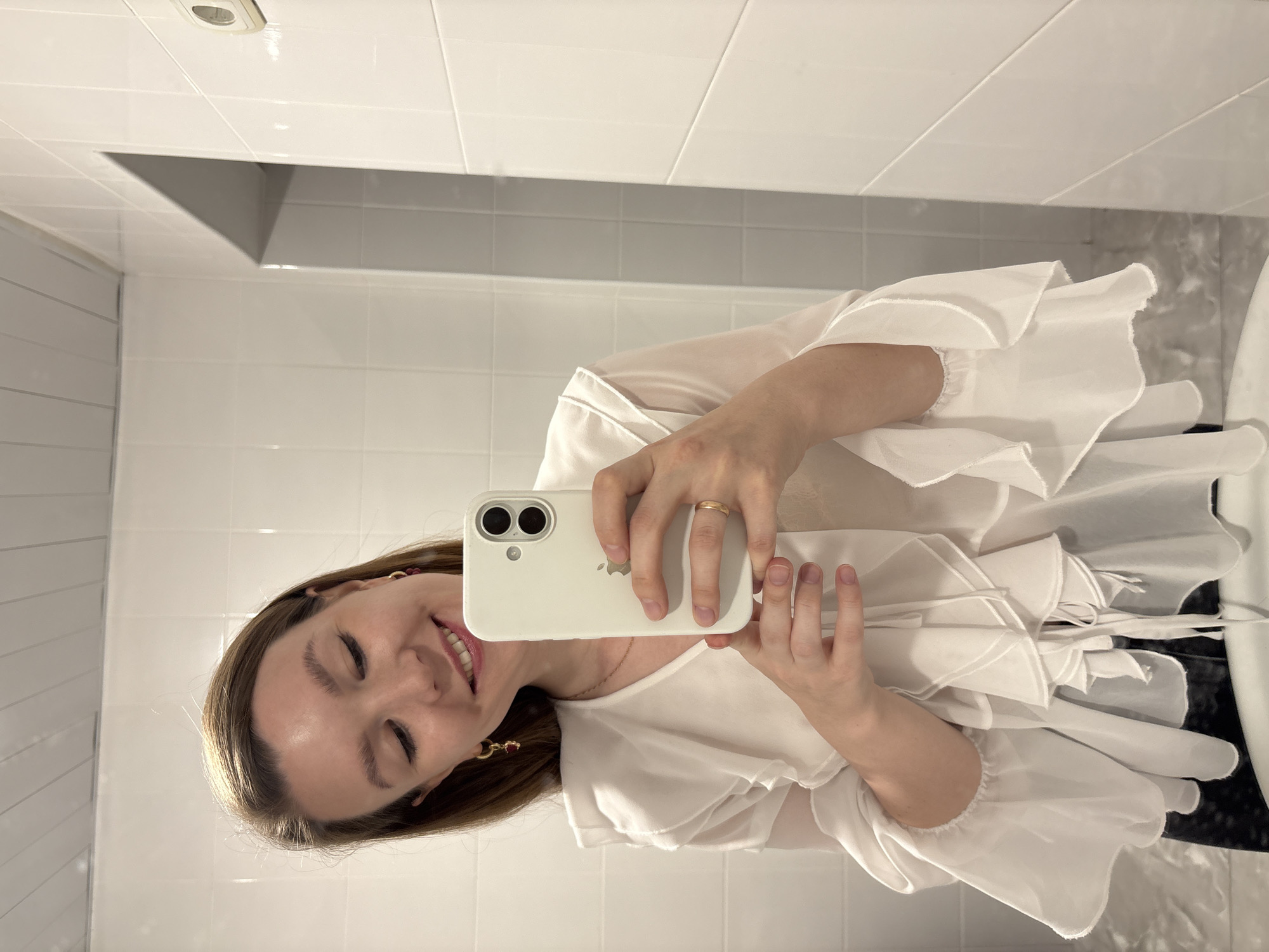 A woman is taking a selfie in a bathroom. She is holding a white smartphone with both hands, showing the screen and the back of the phone to the camera. The woman is smiling, and her face is visible. She is wearing a white ruffled blouse, a gold ring, and earrings. The background is a bathroom with white tiles on the walls and a recessed shelf. This cannot be confirmed as Izhevsk, Russia.