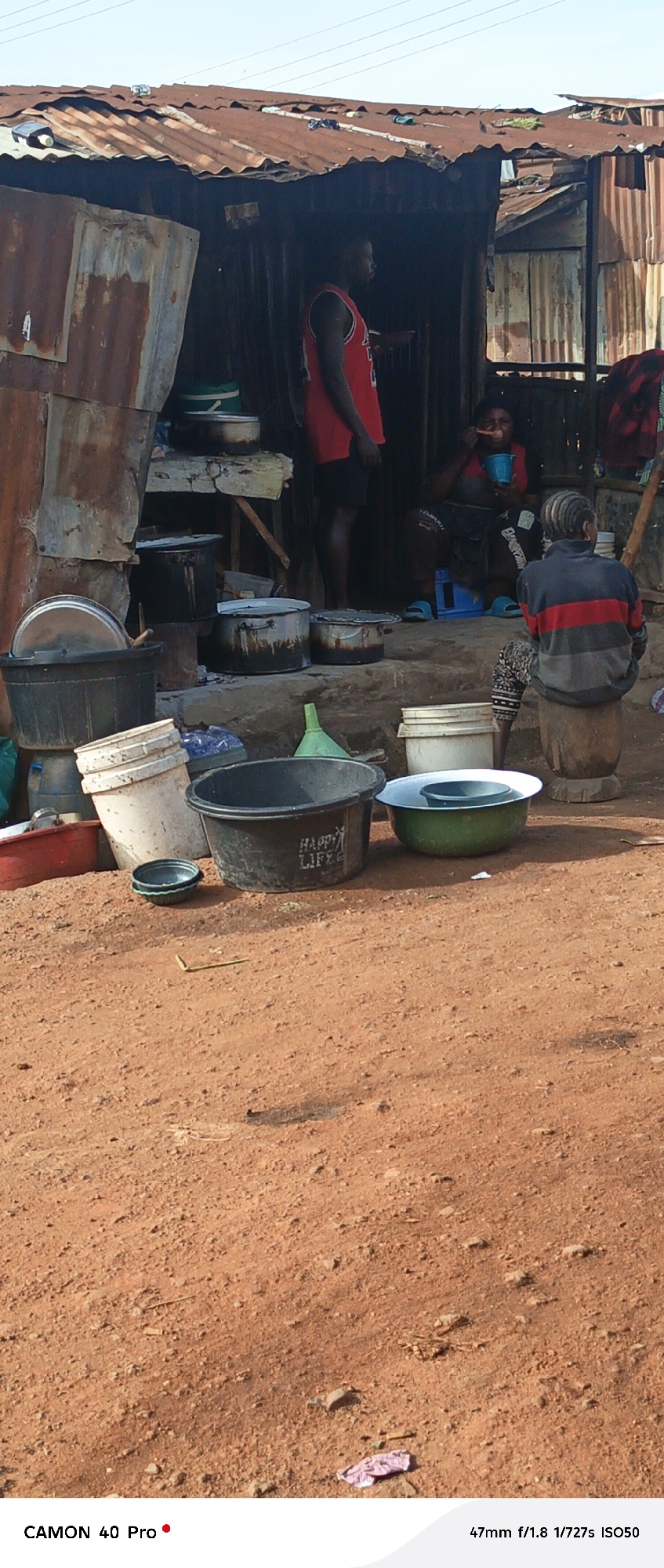 The image depicts an outdoor scene in Barikin Gidar Kanat, Nigeria, with a makeshift shelter constructed from corrugated metal. A man in a red basketball jersey stands near the entrance of the shelter. Inside, a woman is seated and appears to be eating. Another person, viewed from behind, sits on a wooden stool. Various cooking pots, buckets, and basins are placed on the ground and on a raised surface in front of the shelter. The ground is covered in reddish soil with scattered debris. The overall impression is one of a humble, perhaps rural or semi-urban, living environment.