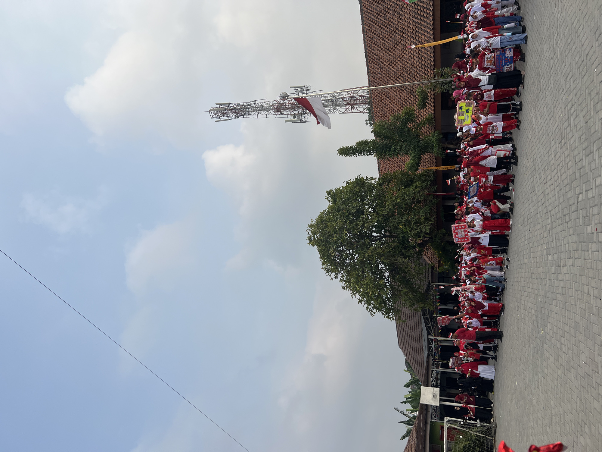 This image depicts an outdoor gathering of a large group of people, predominantly children dressed in red and white attire, on a paved courtyard. The event appears to be a patriotic celebration, with many individuals holding small Indonesian flags, banners, and signs. The children are arranged in rows, facing a direction that is not fully visible in the frame, suggesting a procession or assembly.
In the background, a cluster of buildings with terracotta-tiled roofs are visible, some with open-air covered areas. A tall telecommunications tower stands prominently against a cloudy sky. A large tree with green foliage is positioned between the buildings and the gathering. To the left, a soccer goal is visible, indicating the location might be a school or a sports facility. The ground is paved with interlocking gray bricks.
The presence of Indonesian flags and the red and white theme strongly suggests the location is Indonesia, likely during a national holiday. Given the architectural style and the surrounding vegetation, Gelam Jaya, Indonesia, is a plausible setting for such a scene. The lighting and the overcast sky suggest it is daytime.