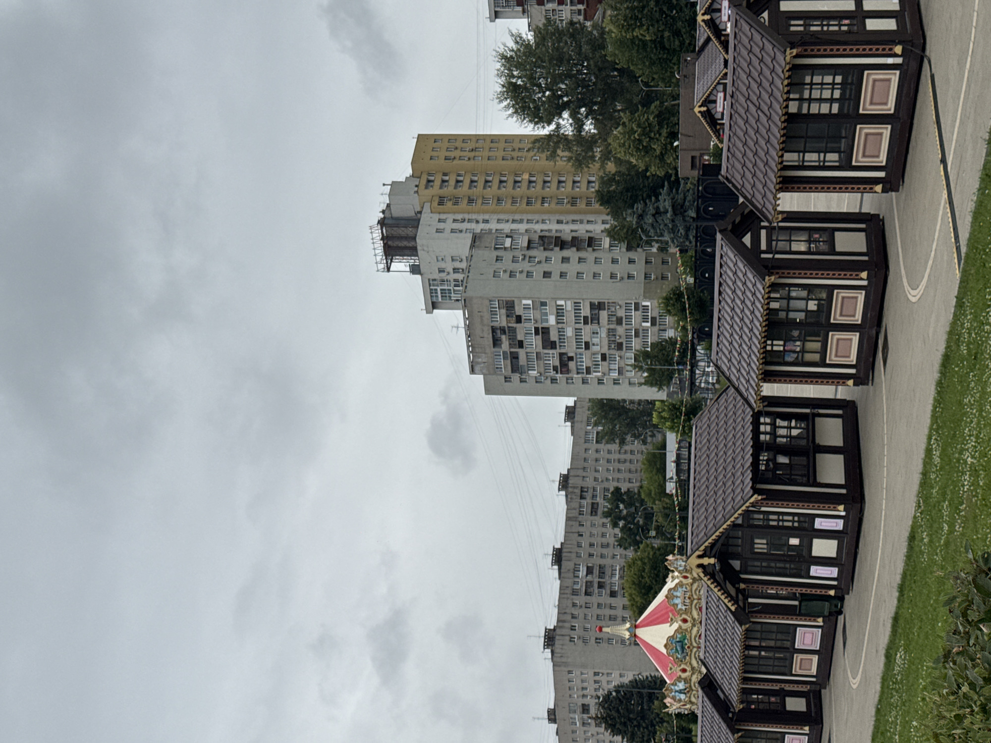 The image shows a line of small, dark brown market stalls with decorative trim and tiled roofs, arranged in a semi-circle on a paved area. Behind the stalls, a colorful carousel with a red and white striped top is visible, along with lush green trees. In the background, several large, multi-story apartment buildings, typical of Soviet-era architecture, dominate the skyline under a cloudy, overcast sky. The setting appears to be a public space within the city of Nizhny Novgorod, Russia, likely a park or square during an event or fair. There are no people visible in the image.