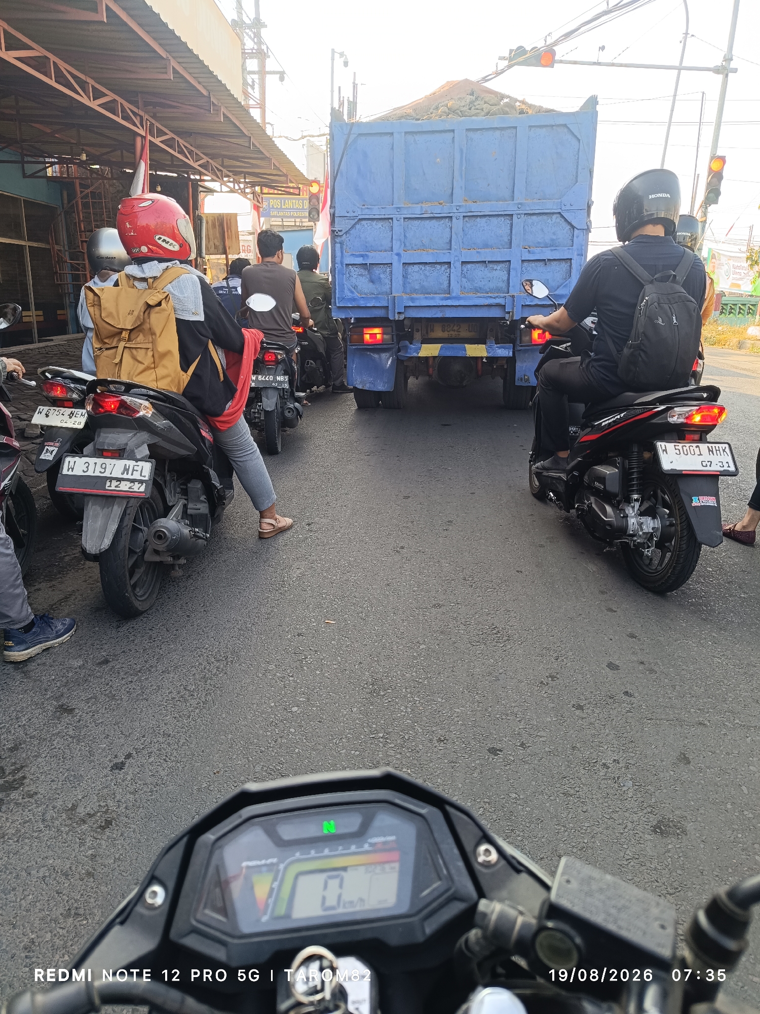 The image depicts a street scene in Sukodono, Indonesia, likely during daylight hours, with the sun casting a warm glow. Several motorcycles and a blue truck are visible on a paved road, suggesting a traffic situation.

In the foreground, the dashboard of a motorcycle is prominently displayed, showing a digital display with "N" for neutral illuminated green, a speedometer indicating 0 km/h, and what appears to be a fuel gauge. The timestamp "19/08/2026 07:35" and "REDMI NOTE 12 PRO 5G | TAROM82" are overlaid on the image.

Behind the foreground motorcycle, a rider wearing a red helmet and a tan backpack is on a black motorcycle. Next to this rider is another person on a motorcycle, and further back, a person in a black shirt with a black backpack is also on a motorcycle. All appear to be stopped, possibly in traffic.

A large blue truck is positioned in the center of the frame, loaded with what looks like dirt or gravel. The truck has Indonesian license plates that read "W 8842 UU". Traffic lights are visible, with one displaying an orange light, indicating a potential stop or caution.

On the right side of the road, there's a building with a corrugated metal roof and some signage, including what appears to be a "POS LANTAS DI SATLANTAS POLRESTO" sign. The overall environment suggests an urban or semi-urban setting. The presence of multiple motorcycles and the truck implies typical street traffic in Indonesia.