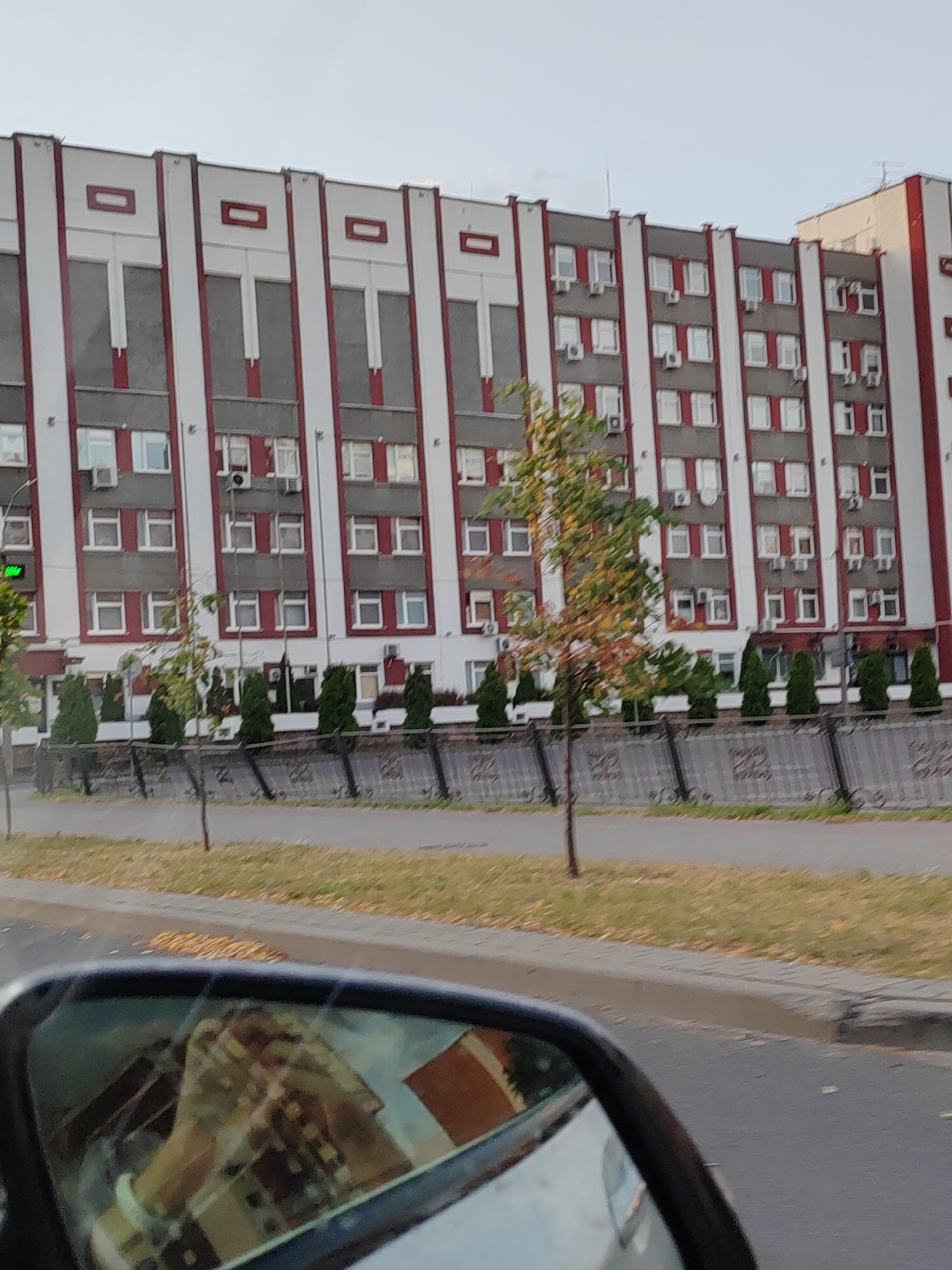 The image shows a large, multi-story building with a facade of gray panels bordered by white and red vertical stripes. The windows are arranged in a grid pattern, with some windows featuring air conditioning units. In front of the building, there is a sidewalk and a street, with a metal fence separating the sidewalk from the building. Several small evergreen trees are planted along the fence. A car's side mirror is visible in the foreground, reflecting a person's hands holding a camera or phone. The setting appears to be a street in the city of Brest, Belarus, possibly on a cloudy or overcast day.
