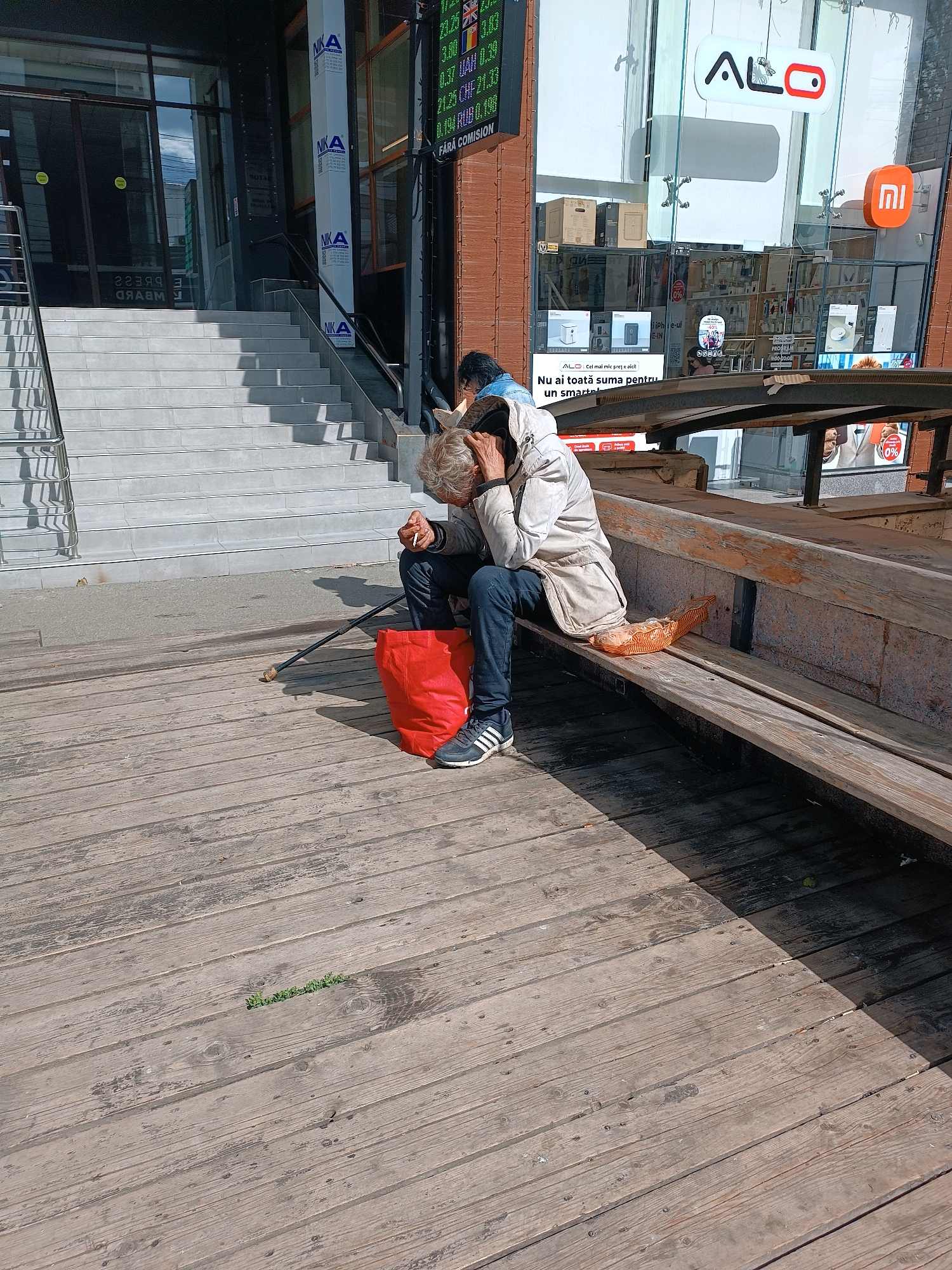 A homeless person sits on a wooden bench outside a storefront, head in hands, holding a cigarette. The person is wearing a light-colored hooded jacket and dark pants, with athletic shoes. To the left, a red bag and a walking stick are placed on the wooden ground. A basket with bread is on the bench next to the person. The storefront features displays of electronic devices and signage in Romanian. A set of stairs leads up to an entrance on the left. The scene appears to be in Chișinău, Moldova, given the Romanian language on the signs.