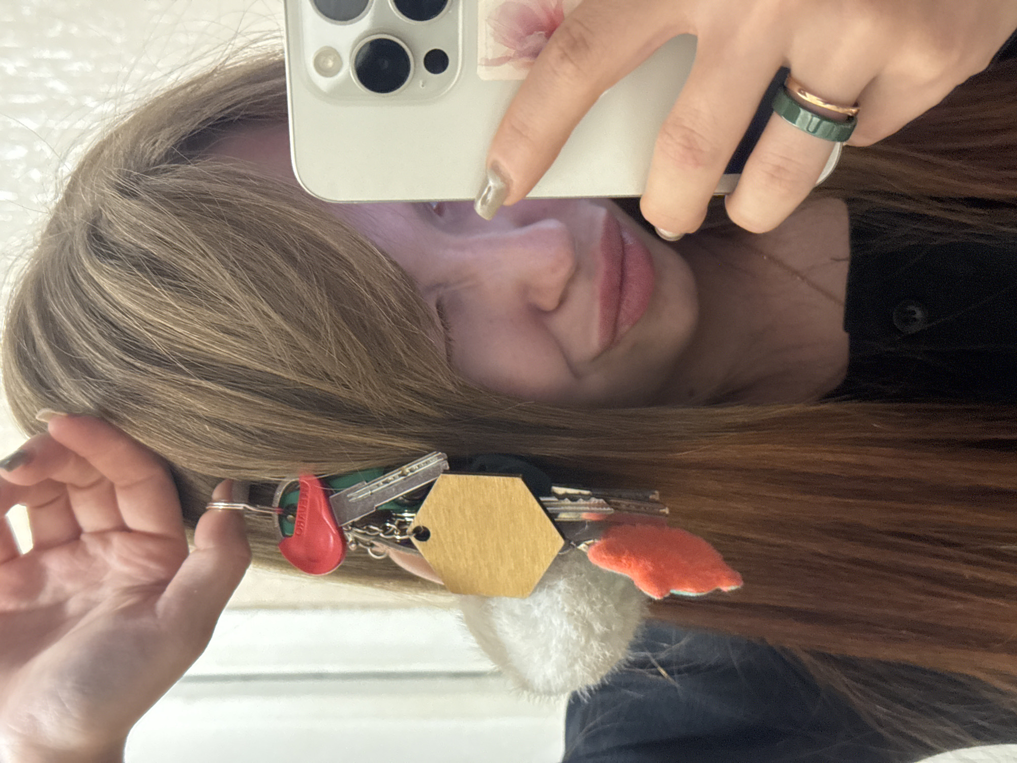 A selfie taken in what appears to be a mirror. The subject is holding a white smartphone, obscuring part of her face. She has long, light brown hair and is wearing rings on her fingers. Attached to her hair near her ear is a set of keys with a red tag, a wooden hexagonal tag, a white fluffy ball, and an orange tag. The location is not discernible from the image.