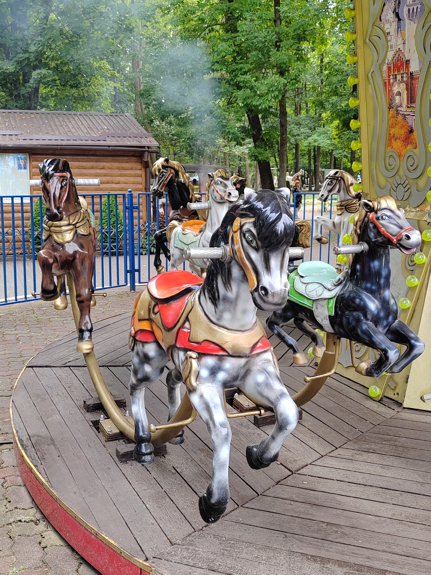 This is a medium-angle, eye-level shot of a carousel at an outdoor amusement park, likely located in Brest, Belarus. The carousel features several painted horses, each mounted on a rocking mechanism. In the foreground, a grey and white speckled horse with a red and gold saddle is prominent. To its right is a black horse with a green saddle. Behind them, a brown horse with a gold saddle is partially visible. In the background, there is a wooden building with a sloped roof, a blue metal fence, and lush green trees. The sky is overcast with a hint of smoke or steam rising from the area. The carousel platform is made of weathered wooden planks. The overall impression is one of a slightly aged but functional amusement ride in a wooded park setting.