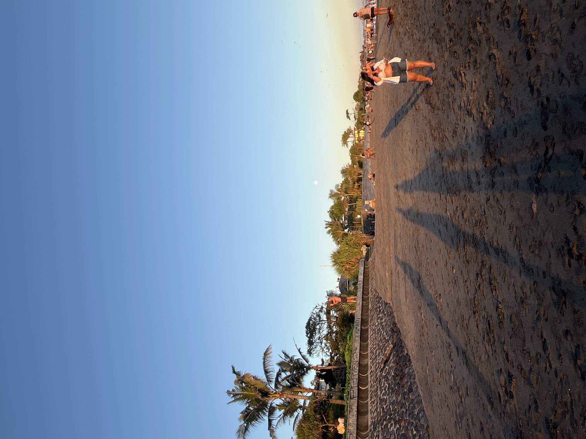 The image shows a beach scene at sunset in North Kuta, Indonesia. Several people are on the dark sand beach. Two people stand close together in the foreground. Palm trees line the shore. The sky is a clear blue. A small moon is visible in the sky. A wall of stones borders the vegetation. The beach has dark sand. Long shadows stretch across the sand from the trees.