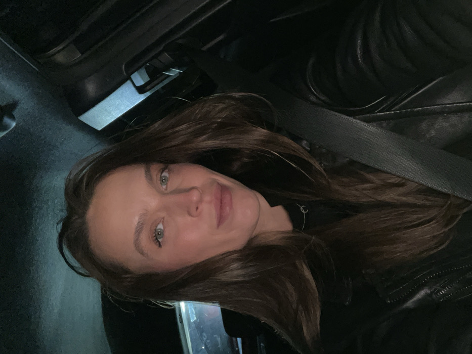 The image displays a portrait of a young woman with long, dark brown hair, fair skin, and striking green eyes, looking directly at the camera with a gentle smile. She is wearing a dark, possibly leather, jacket or top, and a thin silver necklace with a small circular pendant is visible around her neck. A black seatbelt is diagonally stretched across her chest and shoulder, indicating she is seated inside a vehicle.
The setting appears to be the interior of a car, likely at night or in low-light conditions, as the background is predominantly dark and indistinct, showing only vague shapes of the vehicle's interior. The lighting primarily illuminates her face, creating a soft glow. There are no specific visual cues within the image to identify the precise location as Braćeni, Montenegro, other than the general context of being in a car. No text is visible. The mood conveyed is calm and composed.