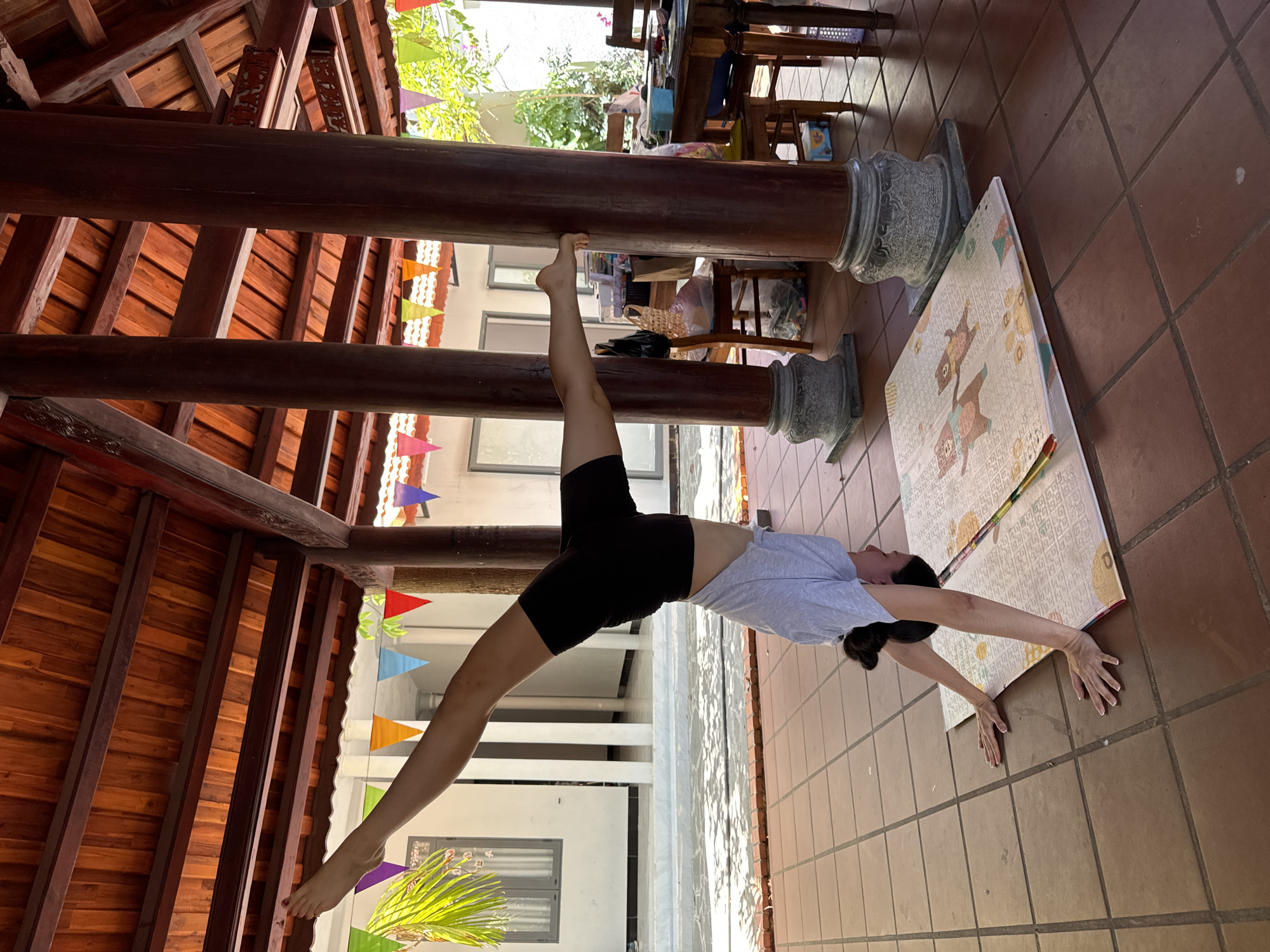 An Asian woman is outdoors, in Mũi Né, Vietnam, doing a gymnastics exercise using a beam or column. She is wearing dark shorts and a light-colored top. She is supported on one hand on a patterned mat on a tile floor, with the other hand raised and legs extended, with one foot secured on a wooden beam of a structure with a dark wood roof. There are decorative flags in the background, and other columns and furniture are visible.