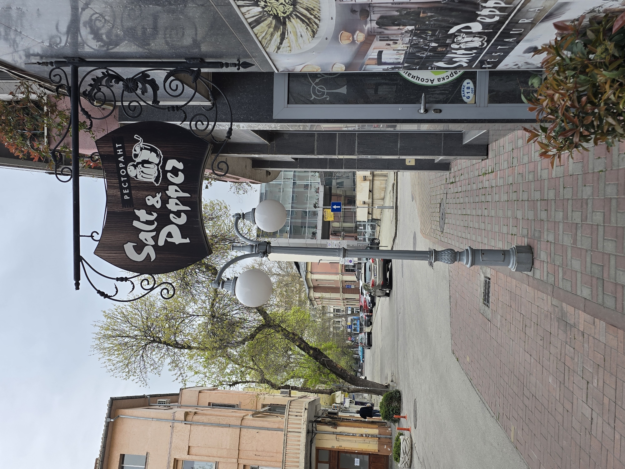 The image depicts a street scene in an urban environment. A restaurant sign, "Salt & Pepper" in Cyrillic script, hangs from an ornate wrought-iron fixture extending from a building on the left. The restaurant's entrance is visible in the background, partially obscured by foliage. A grey street lamp with two spherical lights stands near the sign. The street is paved with red and grey brick, and the buildings lining the street are a mix of architectural styles. Cars are parked along the right side of the street. A person walks in the distance.