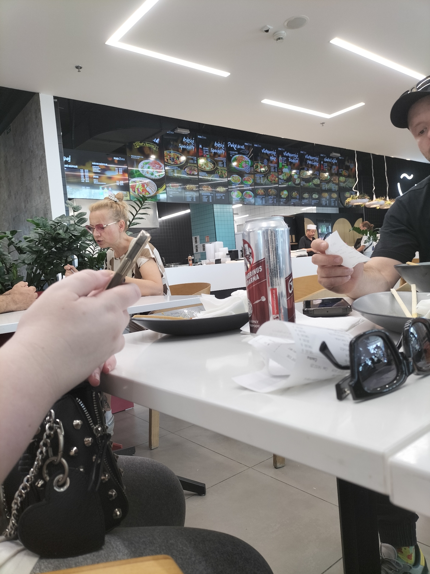 The image depicts a casual dining scene, likely at an Asian food court or restaurant in Pardubice, Czechia. In the foreground, a person's hand holds a smartphone, suggesting they are engaged with their device. A black purse with silver studs and chains is visible, resting on a dark gray chair. On the white table, a can of "Pivní Brina Darma" is prominent, alongside several crumpled receipts and a pair of black sunglasses. Another person, a blonde woman wearing pink-tinted glasses and a striped top, is seated at the table, looking and talking, possibly engaged in conversation or ordering food. Behind her, a large menu board displays various Asian dishes, including "Asijské Poke Bowl," "Pho Levka," and "Vietnamské Way Tradicent." The menu is written in Czech. In the background, other diners and staff members can be seen, with a kitchen area visible behind them. The lighting suggests it is daytime, possibly indoors in a well-lit establishment. The overall atmosphere appears relaxed and informal, characteristic of a food court or casual eatery. The presence of a specific beer brand and Czech language on the menu strongly indicates the location is in the Czech Republic.
