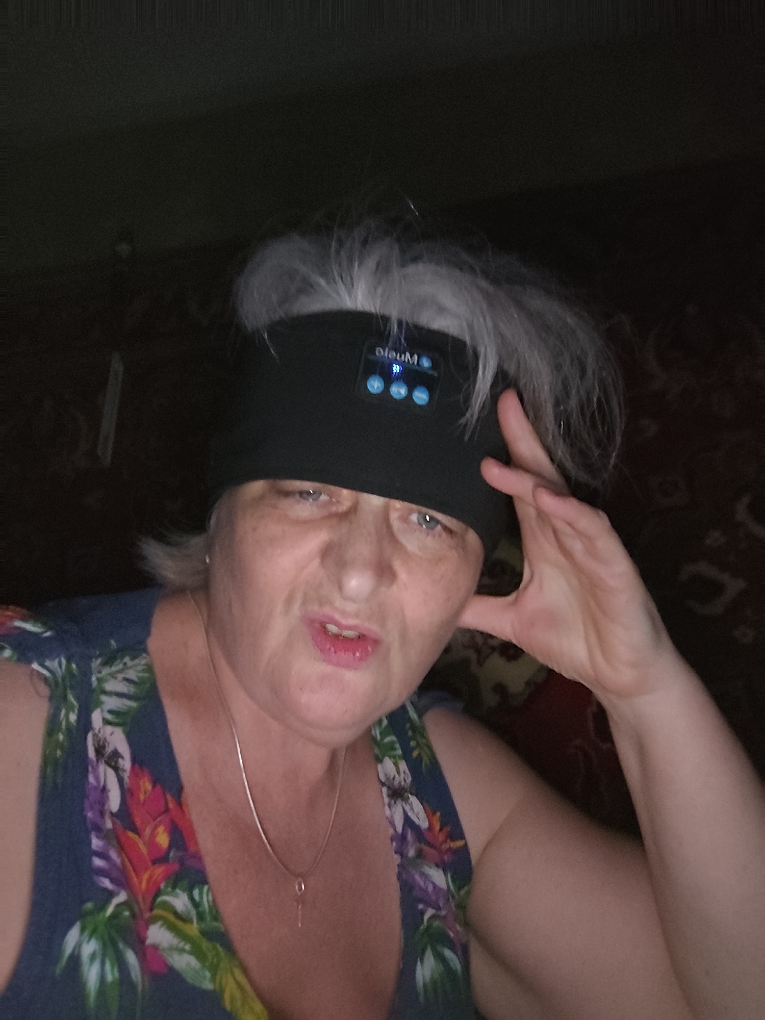 The scene is set in an indoor residential environment within Krasnodar, Russia. A single individual, an adult woman with gray hair, is depicted from the chest up, looking directly towards the camera. She has a slightly parted mouth and a somewhat quizzical or smirking expression. Her right hand is raised, with fingers slightly curled, resting near the right side of her head. She is wearing a dark blue top adorned with a vibrant tropical floral pattern featuring red, orange, purple, white, and green foliage. A thin, silver-colored chain necklace with a small, simple pendant is visible around her neck. A black fabric headband is worn across her forehead and over her hair. Integrated into the front of the headband is an electronic control panel with blue illuminated icons, including the word "OleuM" (or similar inverted text) and several circular buttons, suggesting it is an audio device. The background is dimly lit and consists of a patterned textile, possibly a curtain or tapestry, in dark, muted reddish-brown tones.