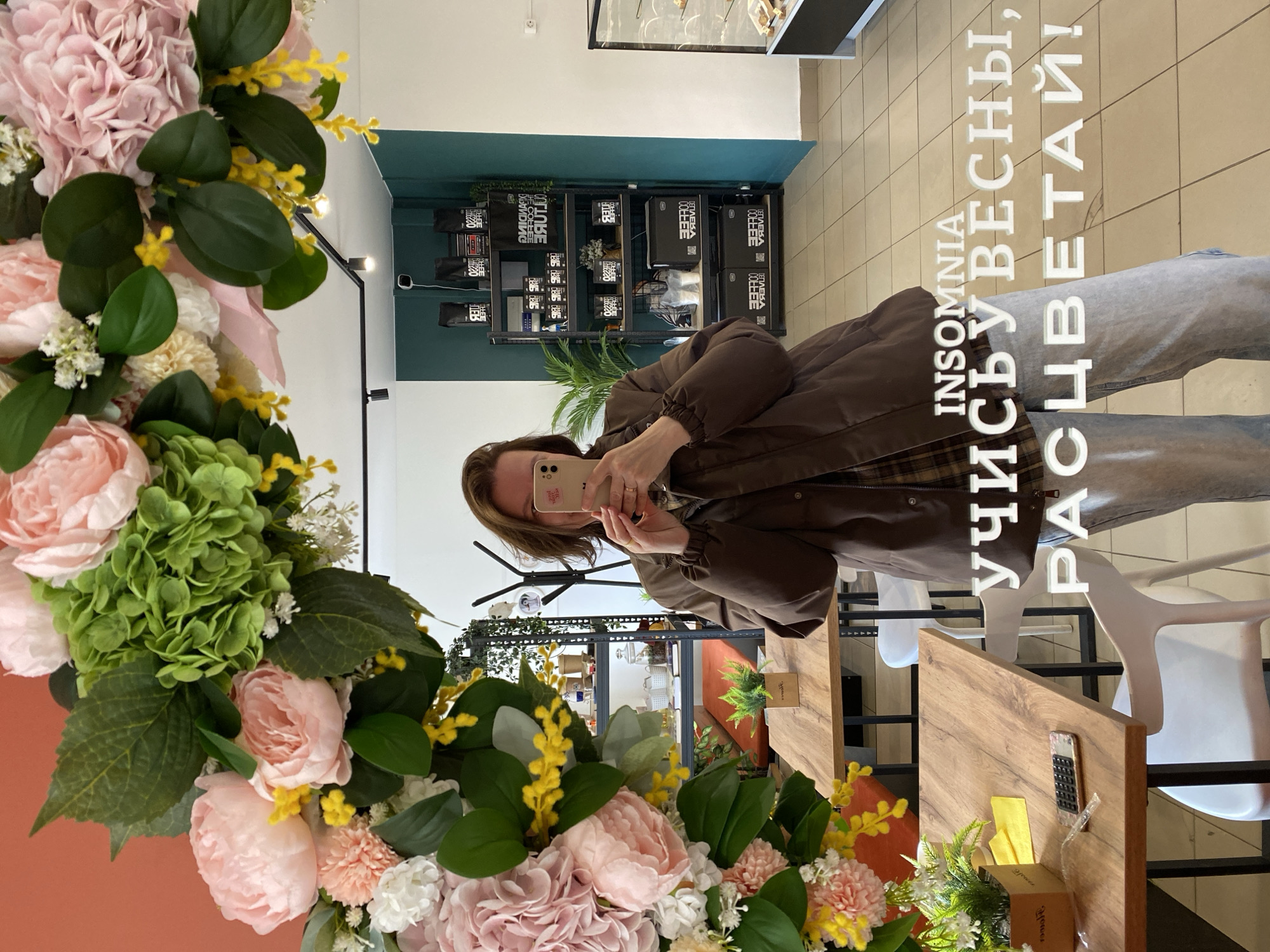 A woman is taking a selfie in a mirror surrounded by an elaborate floral arrangement, likely indoors. The reflection shows shelves stocked with retail products against a teal and white wall. The floor is tiled. A wooden table with a calculator and other small objects is visible in the foreground. The scene suggests a retail environment, potentially a shop. Text is overlaid on the image in Russian, which translates to "INSOMNIA, study, spring, blossom!" The location is likely Chaikovsky, Russia.