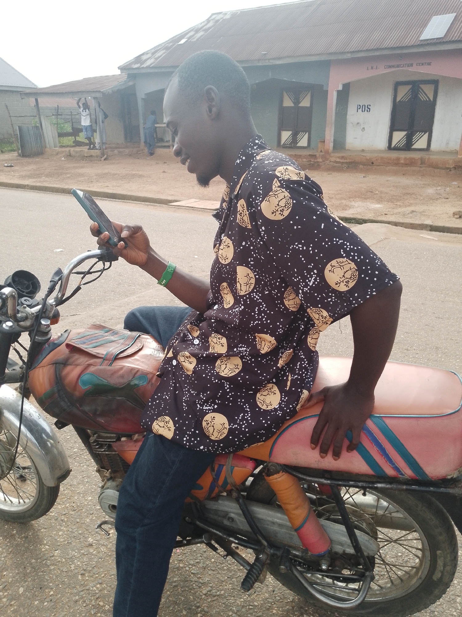 In Balewa, Nigeria, a man sits on a motorcycle in front of a building identified as "A.M.A. - COMMUNICATION CENTRE." The man, wearing a dark shirt with a patterned print and blue jeans, is holding a smartphone in his right hand and appears to be looking at it. A green wristband is visible on his left wrist. The motorcycle has a brown and green patterned seat and visible shock absorbers. In the background, several other individuals are visible near the building, one of whom is standing under a covered area. The environment appears to be an outdoor, possibly dusty, urban setting with a paved road.