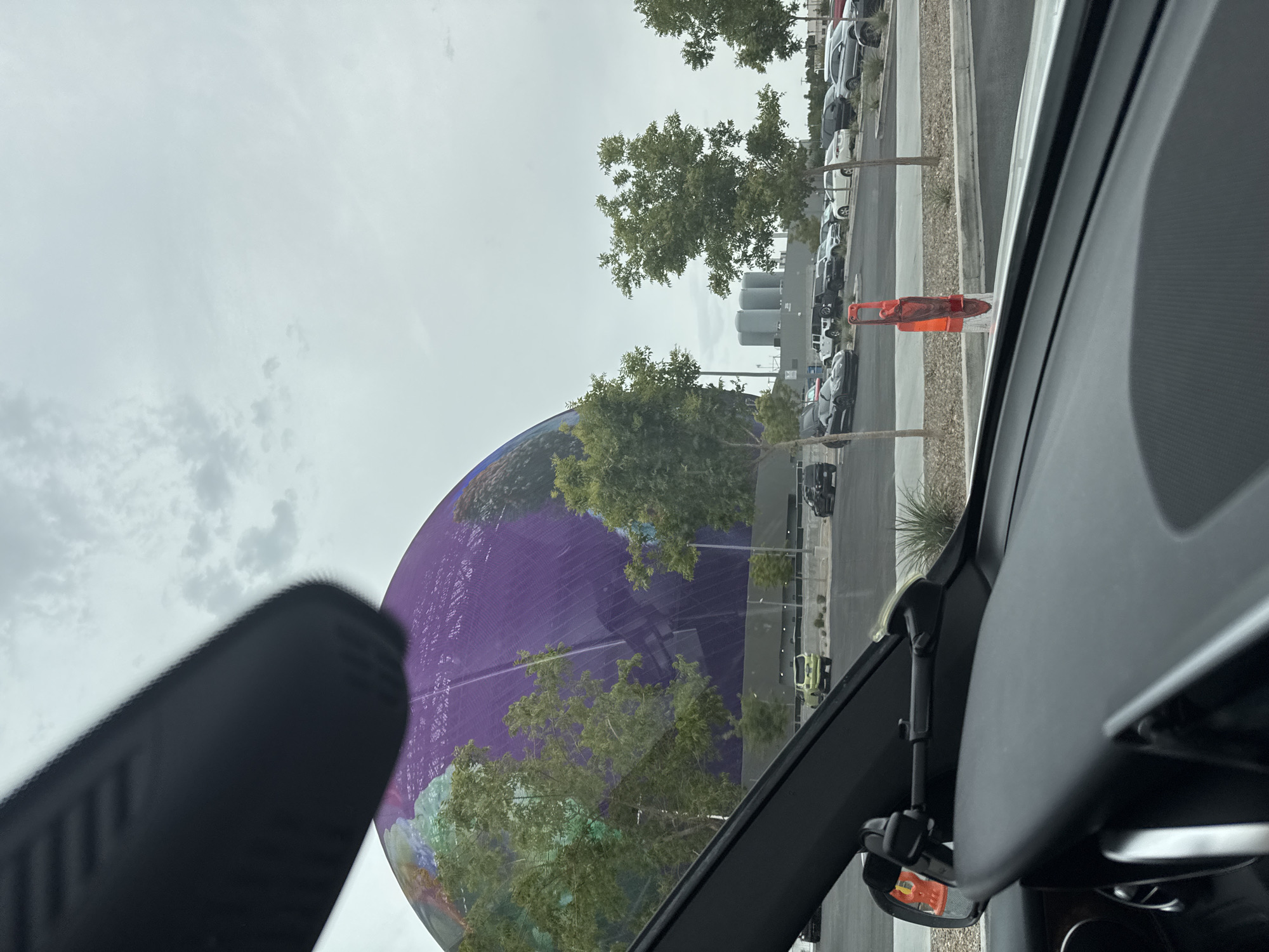 The image shows a view from inside a car, looking out at the Sphere in Paradise, Nevada. The Sphere is a large, spherical building with a colorful, planet-like facade. It is surrounded by trees and a parking lot with several cars. The sky is overcast. A car's rearview mirror and dashboard are visible in the foreground.