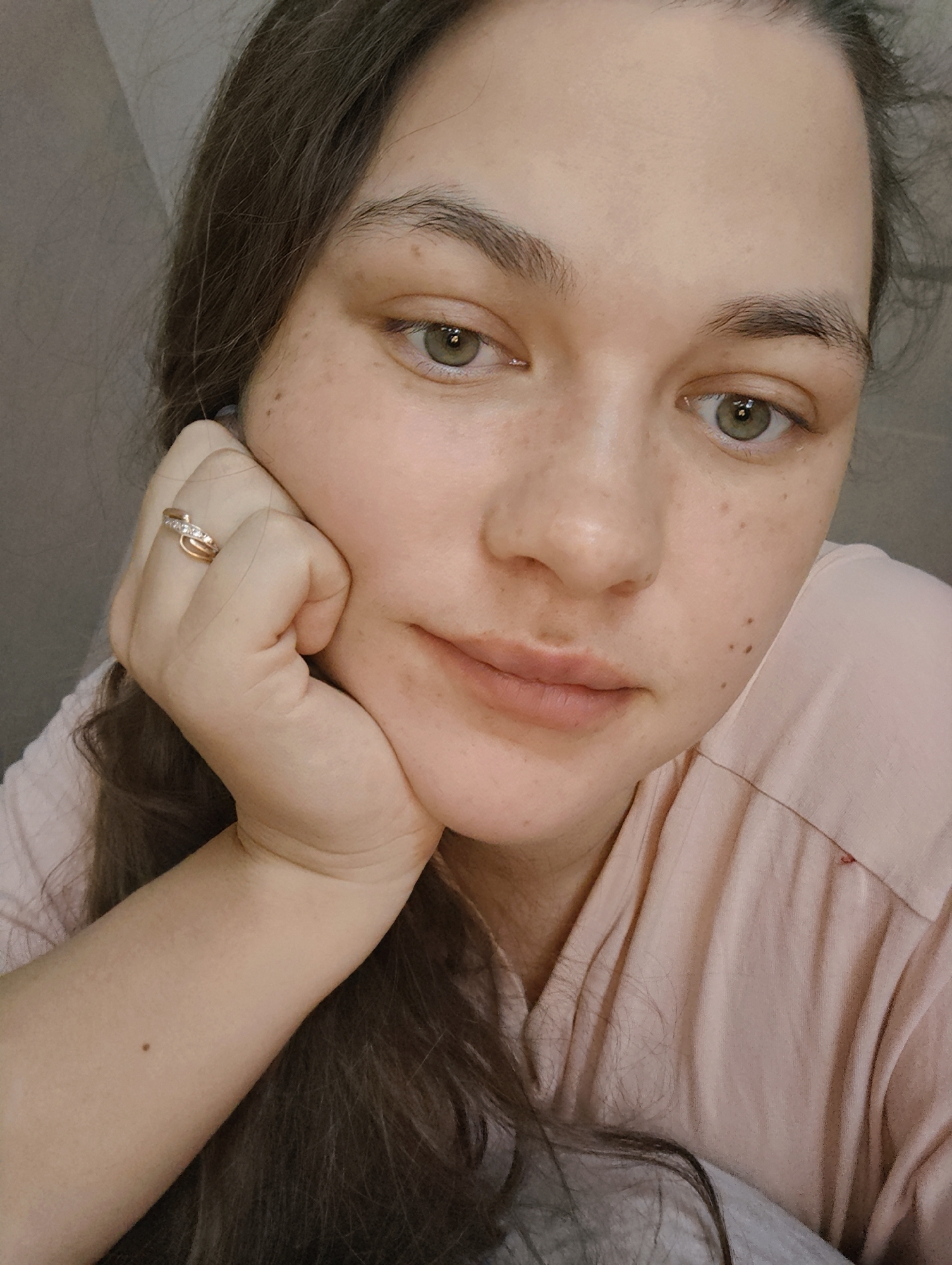 A fair-skinned woman with freckles and green eyes is resting her chin on her hand. She is wearing a gold ring on her finger. Her hair is long and brown. She is wearing a light pink shirt. The background appears to be a neutral wall. The image provides no information about location in the city of Chaikovsky, Russia.