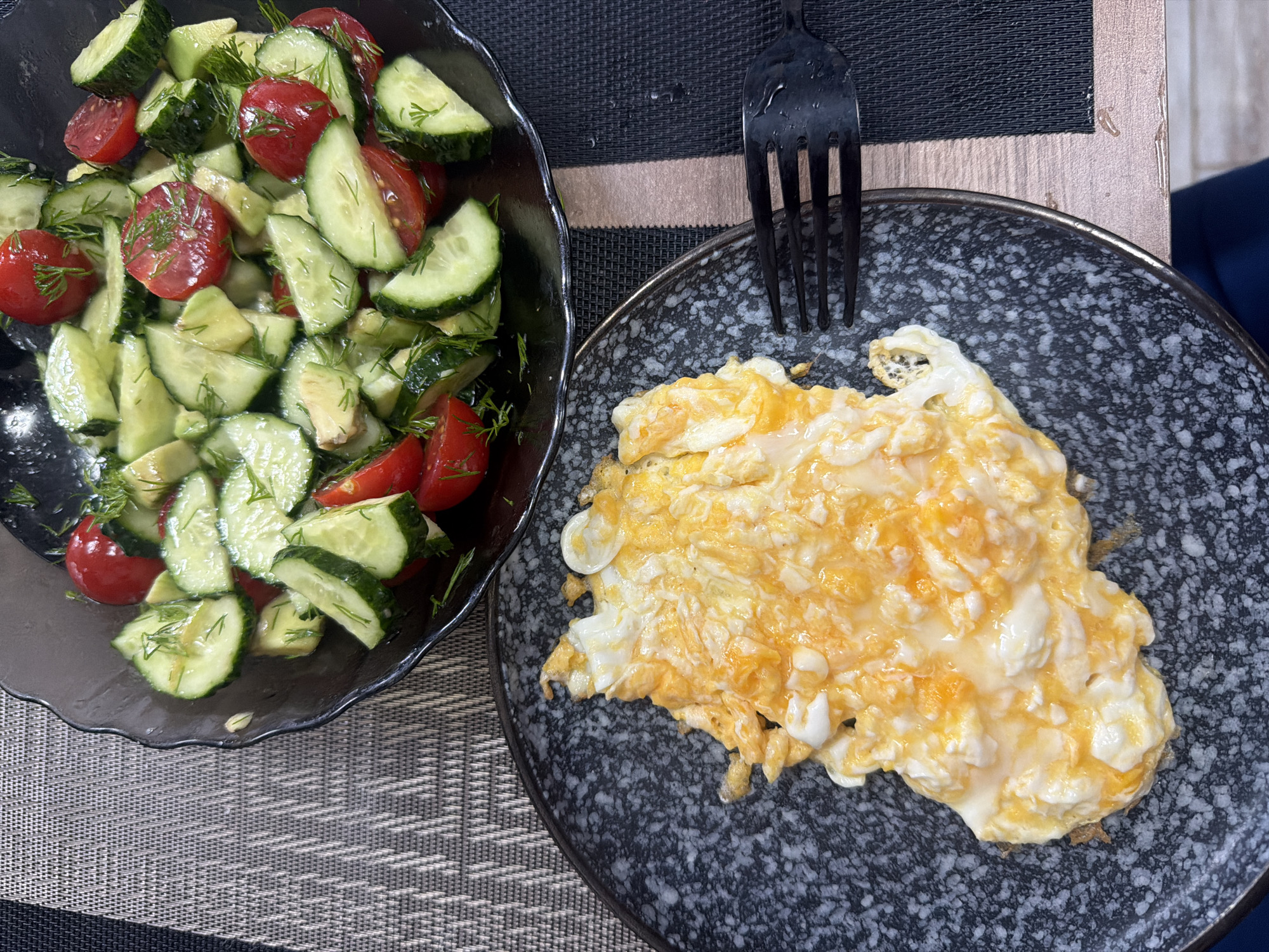 An overhead close-up shot shows a meal of scrambled eggs and a salad. The scrambled eggs are on a dark gray speckled plate, and they are fluffy with visible streaks of white and yellow. A black fork rests to the right of the plate. The salad, in a dark gray bowl with a scalloped edge, consists of sliced cucumbers, halved cherry tomatoes, and chopped dill. The dishes are placed on a striped placemat on a table. The background is blurred. The location is Unknown, Unknown.