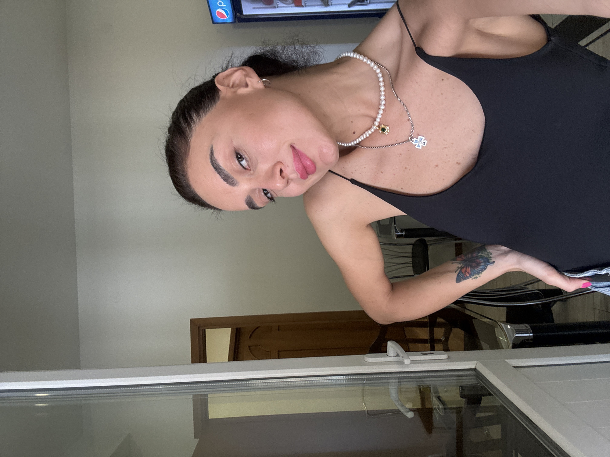A young woman with dark hair pulled back is taking a selfie in what appears to be a domestic setting. She is wearing a black tank top and two necklaces, one with pearls and a green pendant, and another with a silver cross. She has a tattoo of a butterfly on her left arm. In the background, there is a doorway, a refrigerator with a Pepsi logo, and part of a glass door. The environment suggests an indoor location, possibly an apartment or hotel room, in Tbilisi, Georgia.