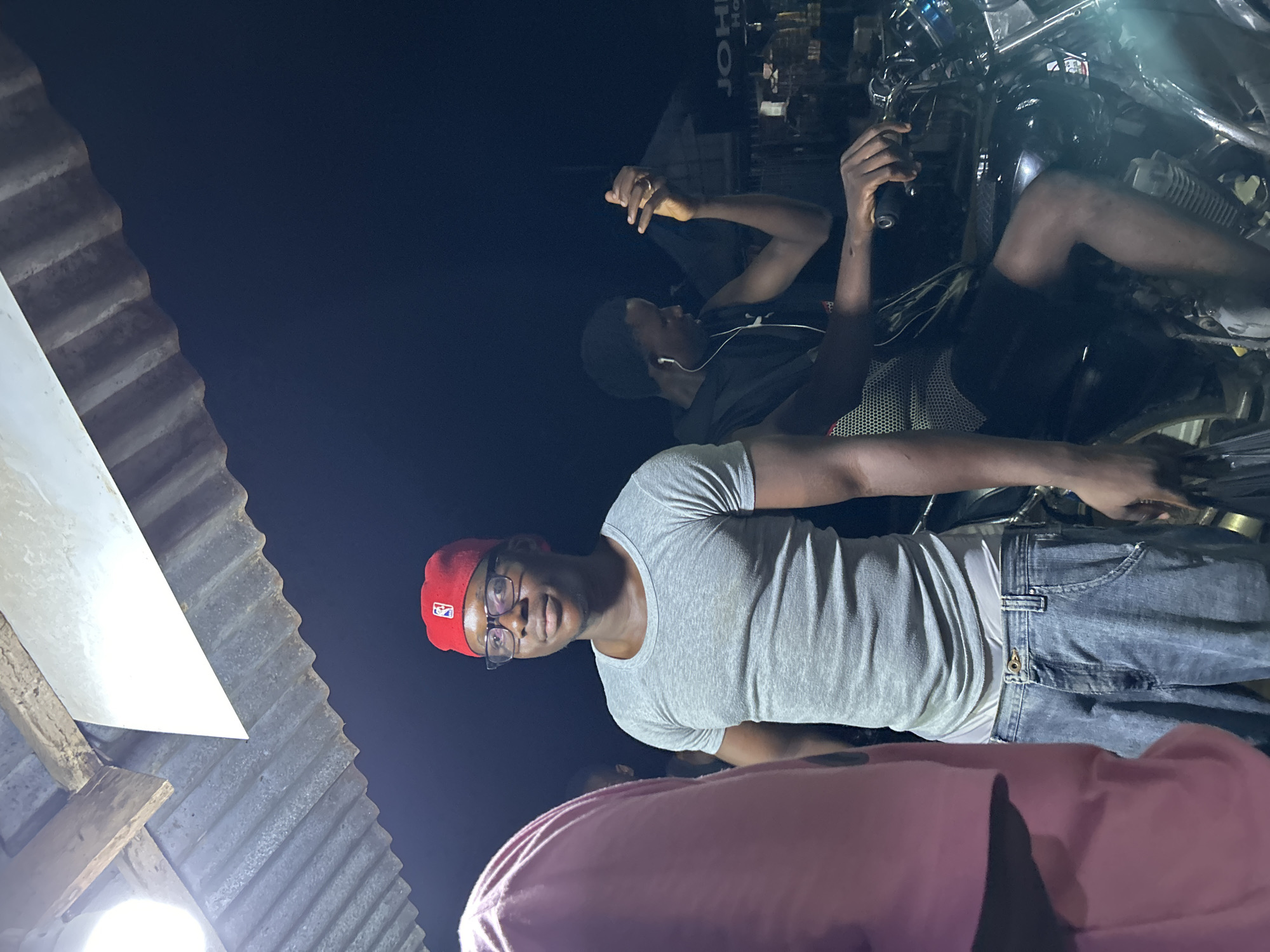 A nighttime scene in Ihiagwa, Nigeria features a man wearing a red cap, eyeglasses, a gray t-shirt, and jeans standing with other people. A motorcycle is visible in the background. An overhead corrugated metal structure is partially shown on the left.