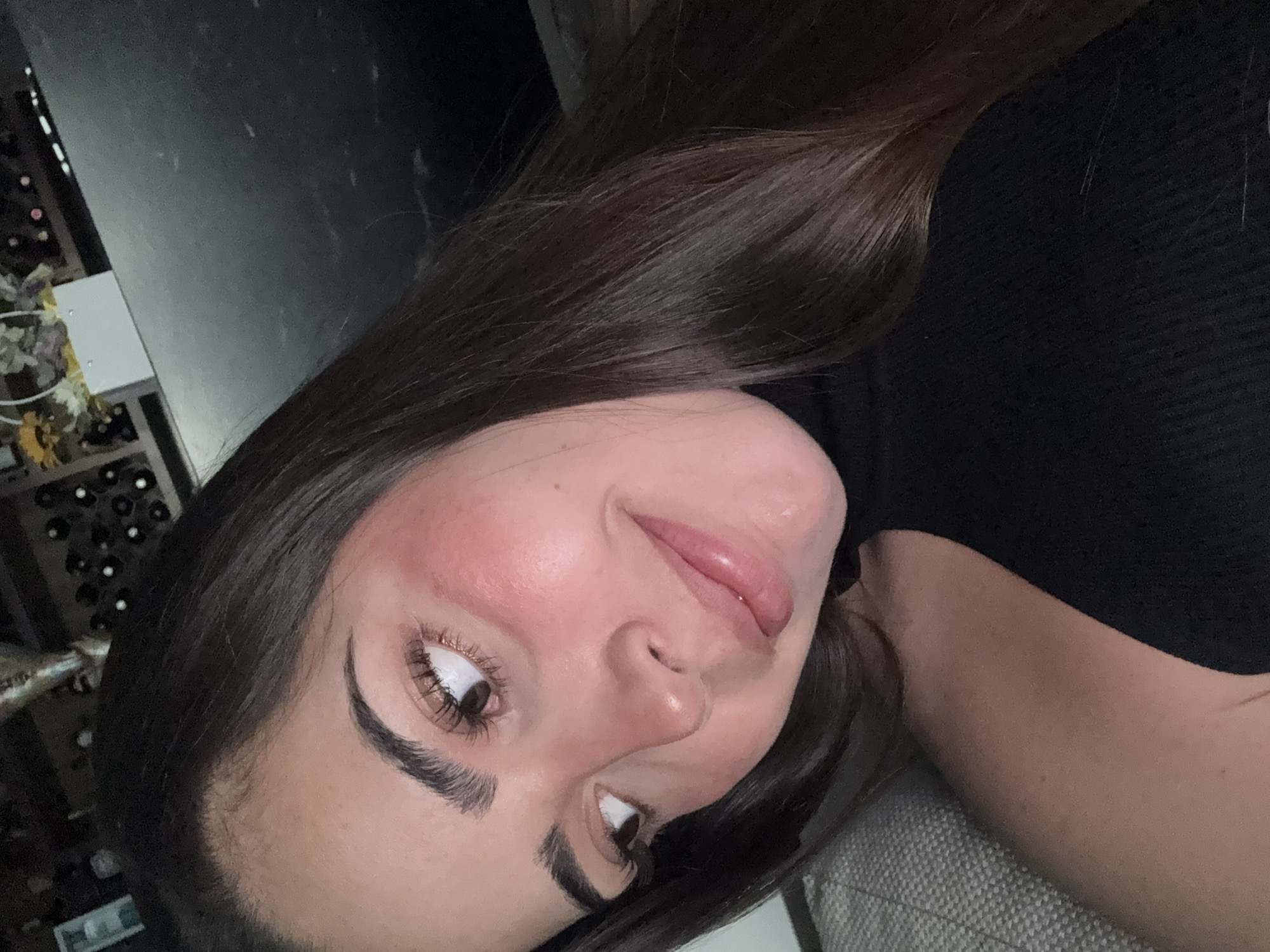 A close-up image depicts a female individual in an indoor setting within San Bernardino Tlaxcalancingo, Mexico. The subject occupies the left side of the frame, showcasing her face from the forehead to the lower jaw, and a portion of her upper torso. She possesses long, dark brown hair, fair skin with noticeable blush on her cheeks, and dark brown eyes directed upward and to the right of the frame. Her eyebrows are dark and well-defined, and her eyelashes appear long. Her lips are full, slightly parted, and exhibit a subtle, glossy smile. She is attired in a black, high-neck, sleeveless top or dress. To her left, a light-colored, textured fabric, possibly upholstery, is visible. The background is dimly lit and out of focus, revealing a structure containing multiple cylindrical objects, consistent with a wine rack, positioned to the upper left. A white rectangular object and light-colored decorative elements are also present in the background, with a dark, flat surface visible on the right.