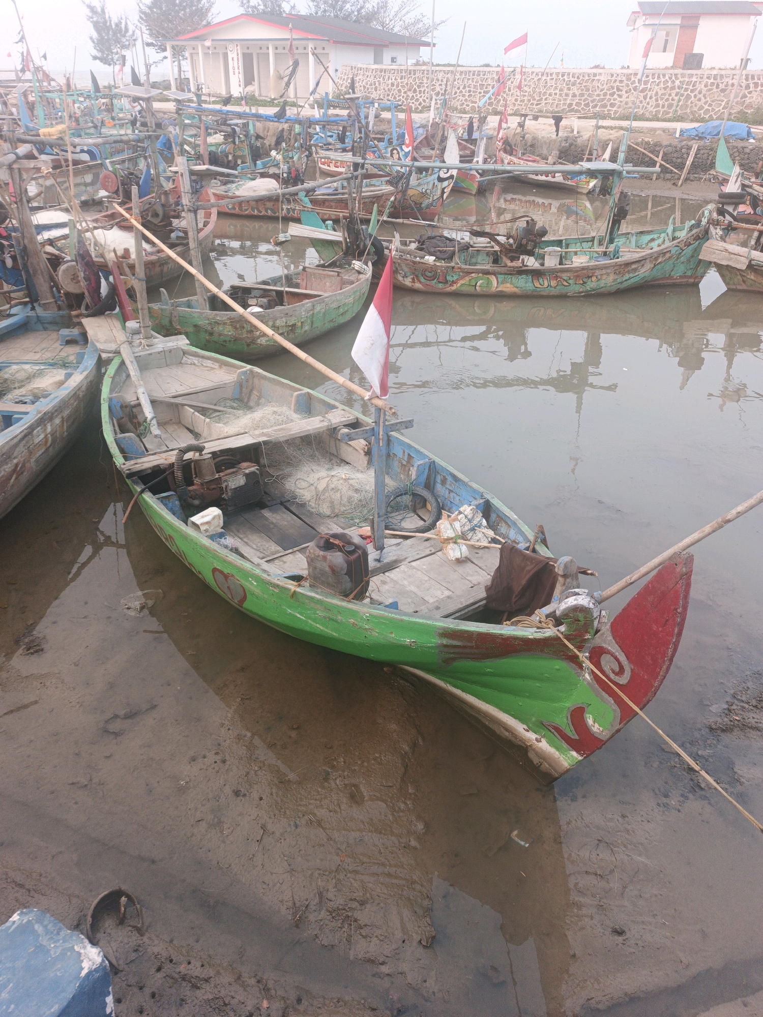The image depicts a harbor in Bumiharjo, Indonesia, filled with numerous fishing boats. The boats are docked in shallow, muddy water. The foreground features a brightly painted green and red fishing boat with an Indonesian flag attached to its stern. The boat contains fishing nets, a small engine, and various containers. In the background, more fishing boats are visible, along with a stone wall and buildings. The sky is overcast, suggesting a hazy or foggy atmosphere. The scene appears to be a typical day at a fishing harbor.