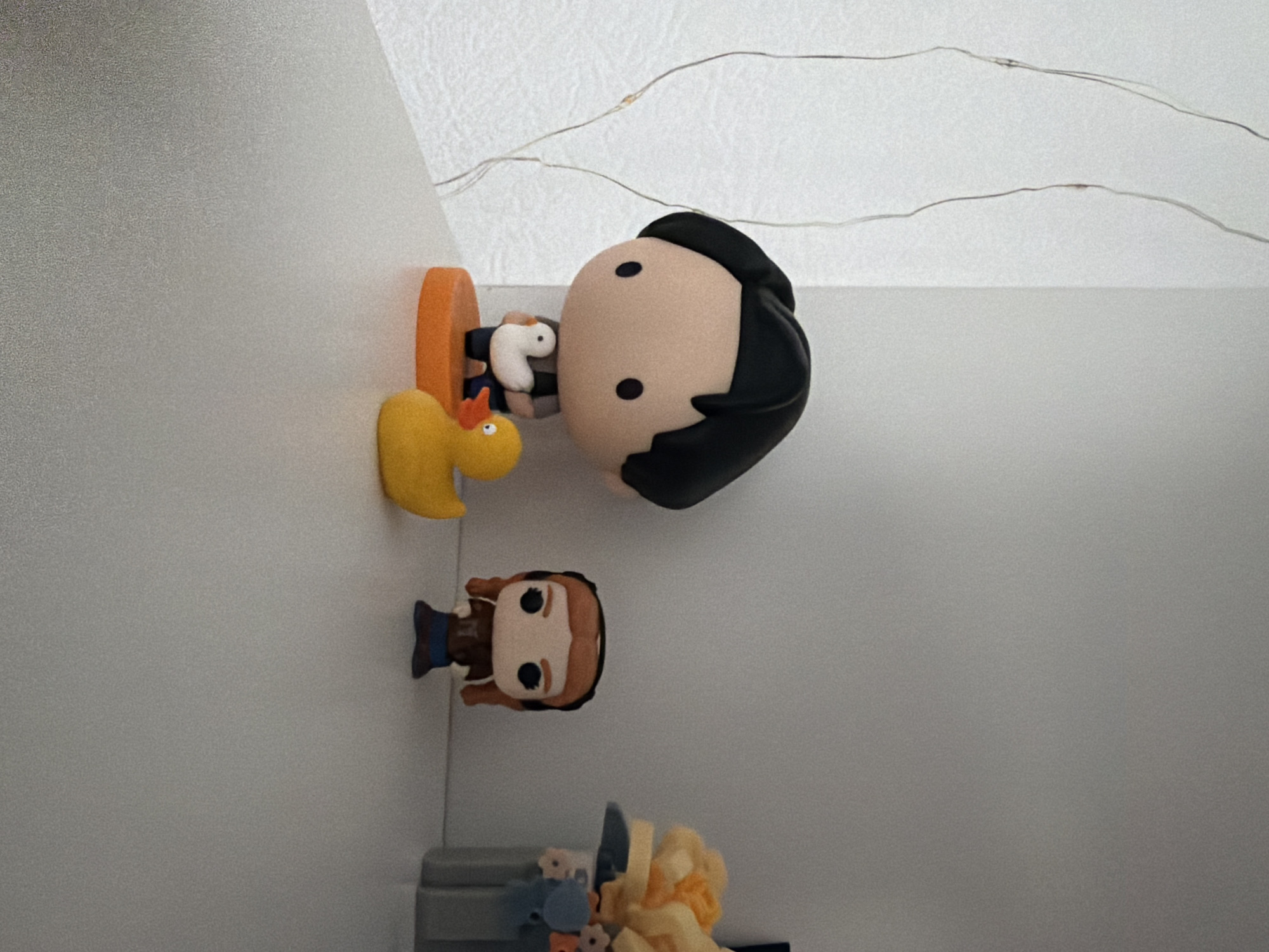 The image depicts a white shelf with several decorative figurines and objects. On the left side of the shelf, there is a large-headed figurine with black hair, wearing a grey shirt and dark pants, holding a small white duck. This figurine stands on an orange circular base. To its right, a yellow rubber duck is positioned. Further to the right, a smaller figurine with brown braided hair, wearing a brown jacket and blue pants, is visible. In the background, on the far right, a grey container holds artificial yellow and orange flowers. The shelf appears to be in an indoor setting, possibly a home or office, with a textured white wall behind it. There are no explicit geographical markers or elements that would indicate the location in Kyiv, Ukraine.