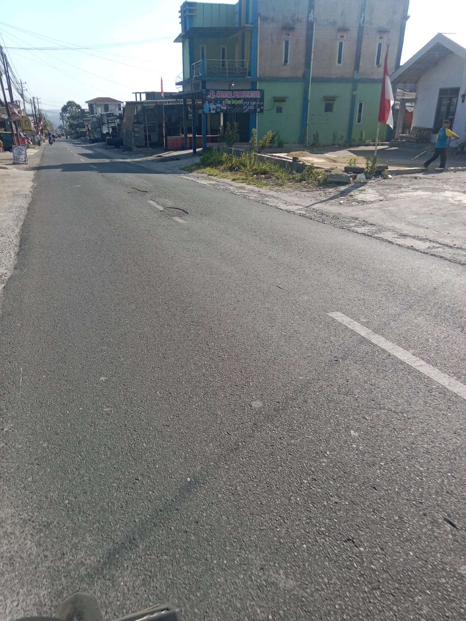 A street scene in Kertek, Indonesia, shows a paved road with a white line marking the lane. On the right side of the road, there is a building with a green and beige facade, and a red and white flag is planted in front of it. A person in a blue and yellow shirt is sweeping the ground with a broom. Further down the road, other buildings and a sign that reads "RENTAL PLAYSTATION THE LEGEND PS3 PS4" are visible. On the left side of the road, there are more buildings, a street light pole, and several motorcycles are parked. The sky is clear and blue, and the sun is shining.