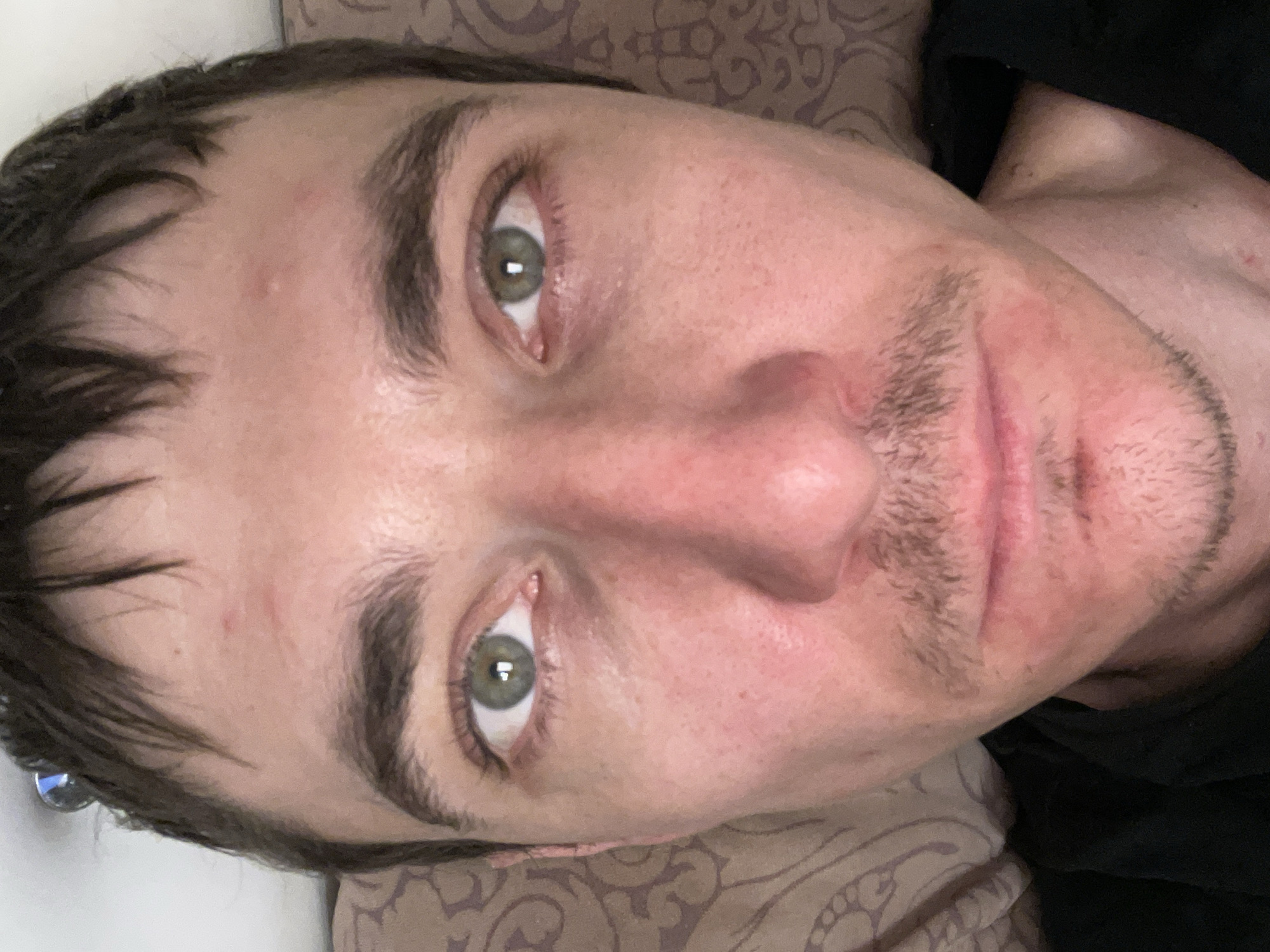 A close-up, overhead shot shows the face of a young man with light skin and dark brown hair. His hair is short and somewhat messy. He has green eyes, dark eyebrows, and light stubble on his upper lip and chin. His face appears flushed and there are some small red marks and blemishes on his forehead, cheeks, and around his mouth. He is wearing a black shirt and lying down, with a pillow with a brown patterned fabric visible behind his head. The image appears to be taken indoors, possibly in a bedroom. There is no indication of the city of Laishevo, Russia within the image itself.