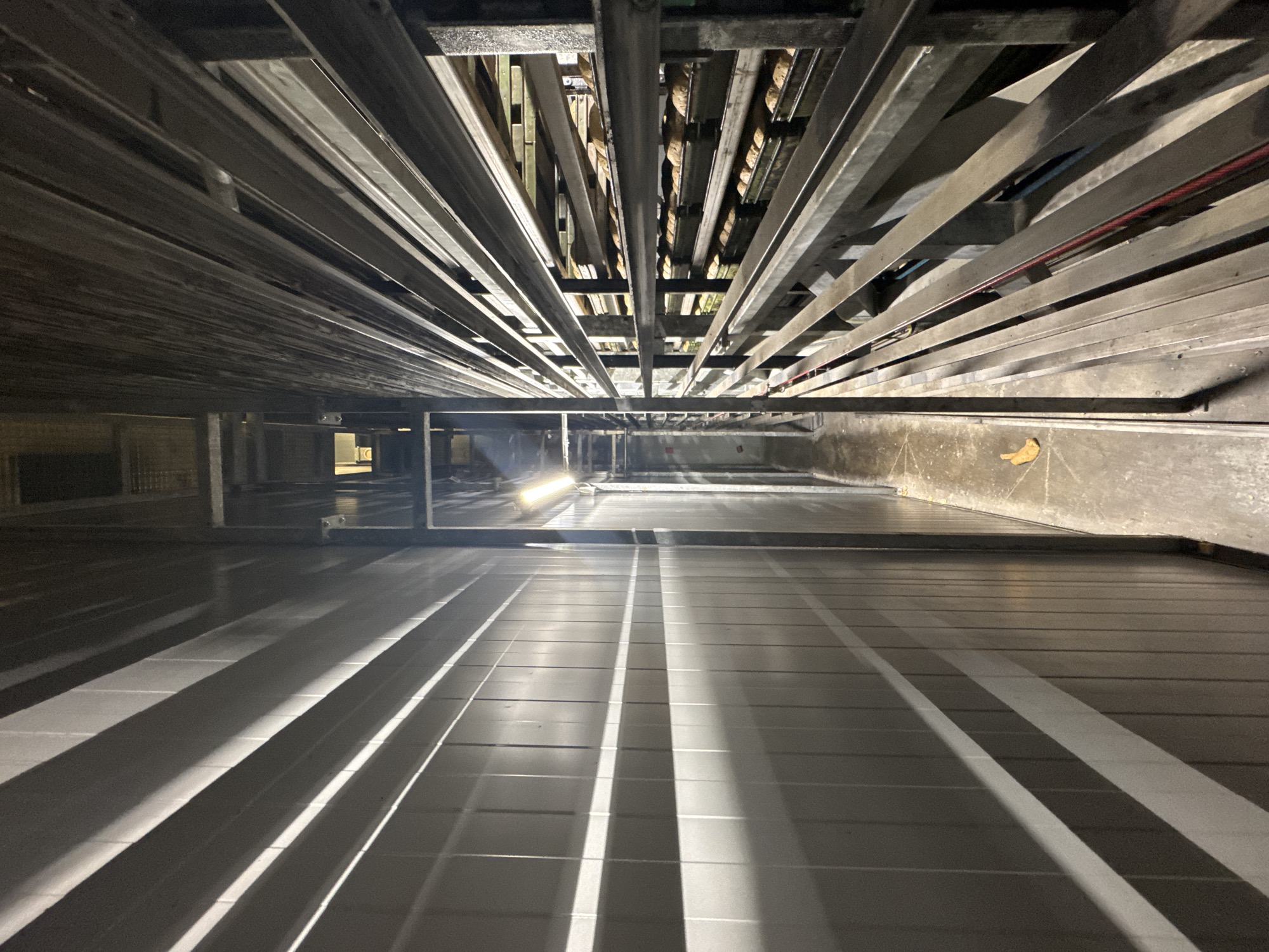 The image depicts the interior of a commercial bakery's bread cooling tunnel or storage area, likely located in Dedovsk, Russia. The scene is characterized by a narrow passageway with a ribbed, light-colored wall on the left and a multi-tiered metal shelving unit on the right. The shelving unit is filled with numerous loaves of bread, some of which are in dark rectangular pans. The lighting is artificial, with a prominent fluorescent light fixture visible on the right side of the passageway. Shadows are cast across the ribbed wall, suggesting a directional light source. The floor appears to be concrete and is somewhat damp in places. A single piece of what appears to be discarded food packaging lies on the floor. The overall impression is one of industrial food production and storage.