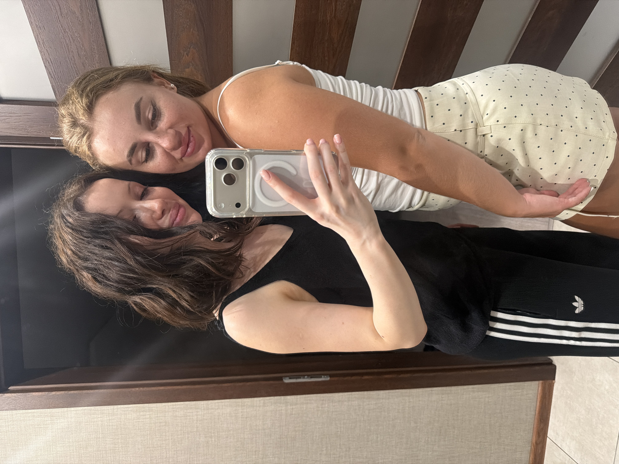 Two young women are taking a selfie in a room that appears to be a hotel or apartment. The woman on the left has long, dark, wavy hair and is wearing a black tank top and black athletic pants with white stripes. The woman on the right has blonde hair pulled back and is wearing a white tank top and a light-colored skirt with black polka dots. They are standing close together, with the woman on the left holding the phone to take the photo. The background shows a doorway with wooden panels and textured wallpaper. The flooring appears to be tiled. The overall setting is domestic or hospitality-related. There is no information in the image to indicate a specific location within the city of Zaporizhzhia, Ukraine.