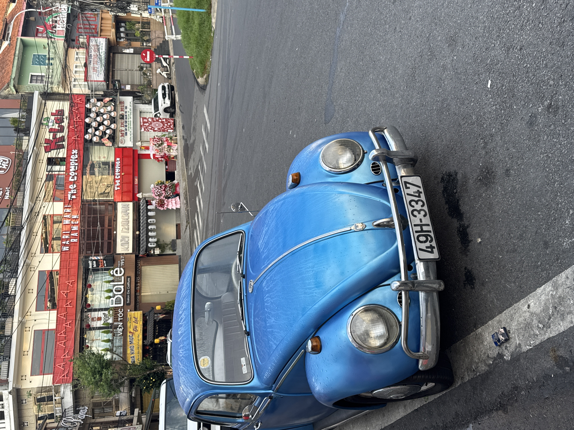 A blue classic Volkswagen Beetle is prominently parked on the side of an urban street in Da Lat, Vietnam. The car, adorned with chrome bumpers and trim, appears to have water droplets on its surface, suggesting recent rain or washing. Its license plate, "49H-3347", is clearly visible.
The street is lined with multi-story commercial buildings, displaying a mix of architectural styles and numerous business signs. Dense overhead electrical and communication wires crisscross the scene. In the background, a white car and a white scooter are also parked.
Visible text on the shop signs includes: "WAI HARI RAMEN", multiple instances of "The Complex", "VIỆN TÓC BaLê" (Hair Salon BaLe), "Salon De Coiffure", "1957", "KIRI IZAKAYA", "MỸ DỤNG HUE" (Hue Beauty Supplies), "CUNG CẤP NỘI THẤT SALON TÓC - NAIL" (Supplies for Hair - Nail Salon Interior), "BIA KHO MỤC", "Da Lat" (confirming the location), and "SABER SHOP MAN'S 78" (possibly "BARBER SHOP MAN'S 78"). There are also generic signs with repeated "AAAAAAA" text and "NAM" and "Lab". Decorative floral arrangements are visible outside some storefronts.
The time of day is daytime, with bright, even lighting. The setting is a bustling street in a commercial district, characteristic of a Vietnamese city. No people are explicitly visible in the frame, but the array of businesses suggests a lively commercial environment.