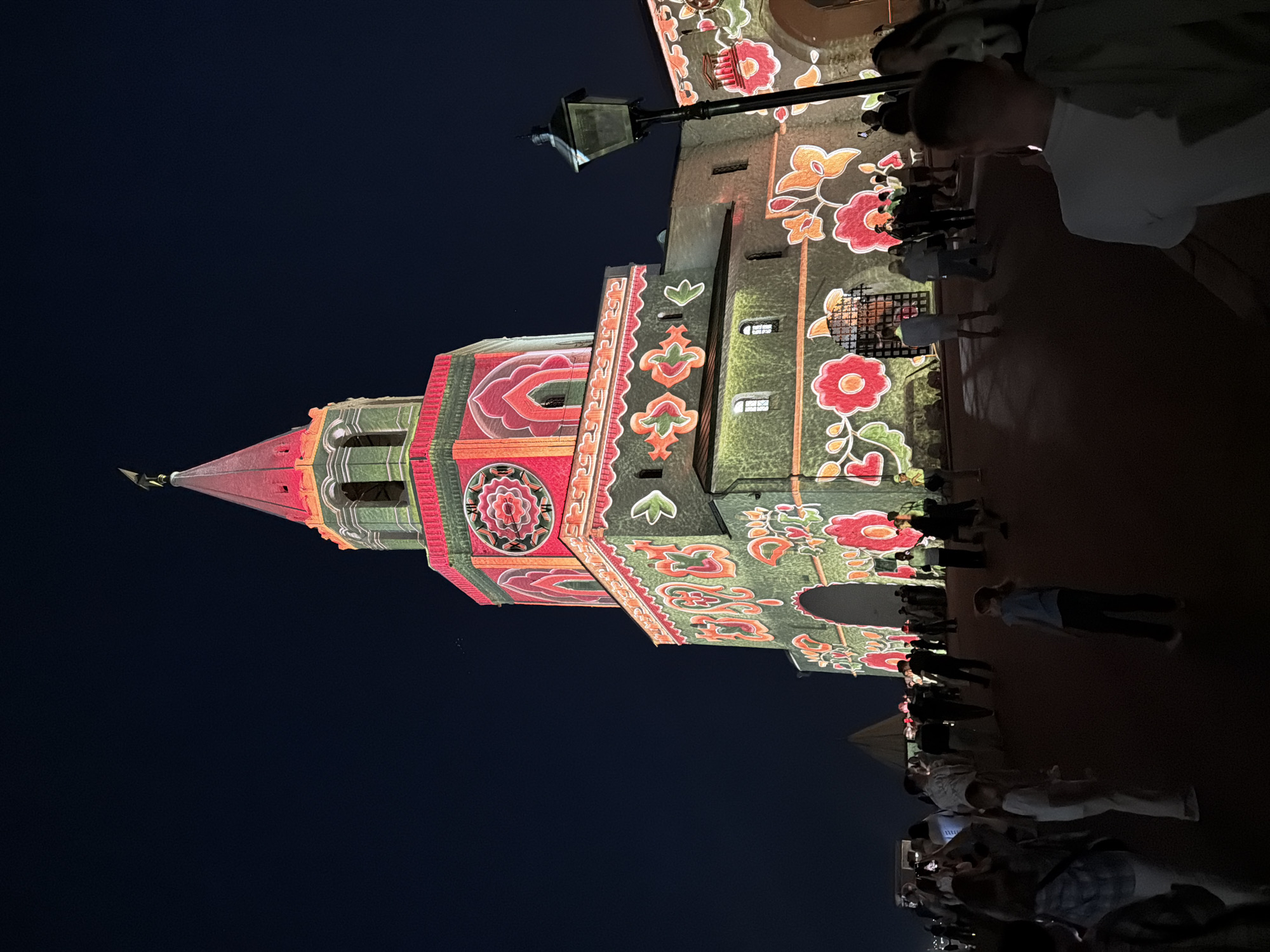 At night, a tall, ornately decorated tower in Kazan, Russia is the focus of a light show. The tower is illuminated with colorful floral patterns in reds, greens, yellows, and oranges. A crowd of people stands facing the tower, their silhouettes visible against the illuminated facade. A lamppost extends from the side of the tower. The sky is dark.