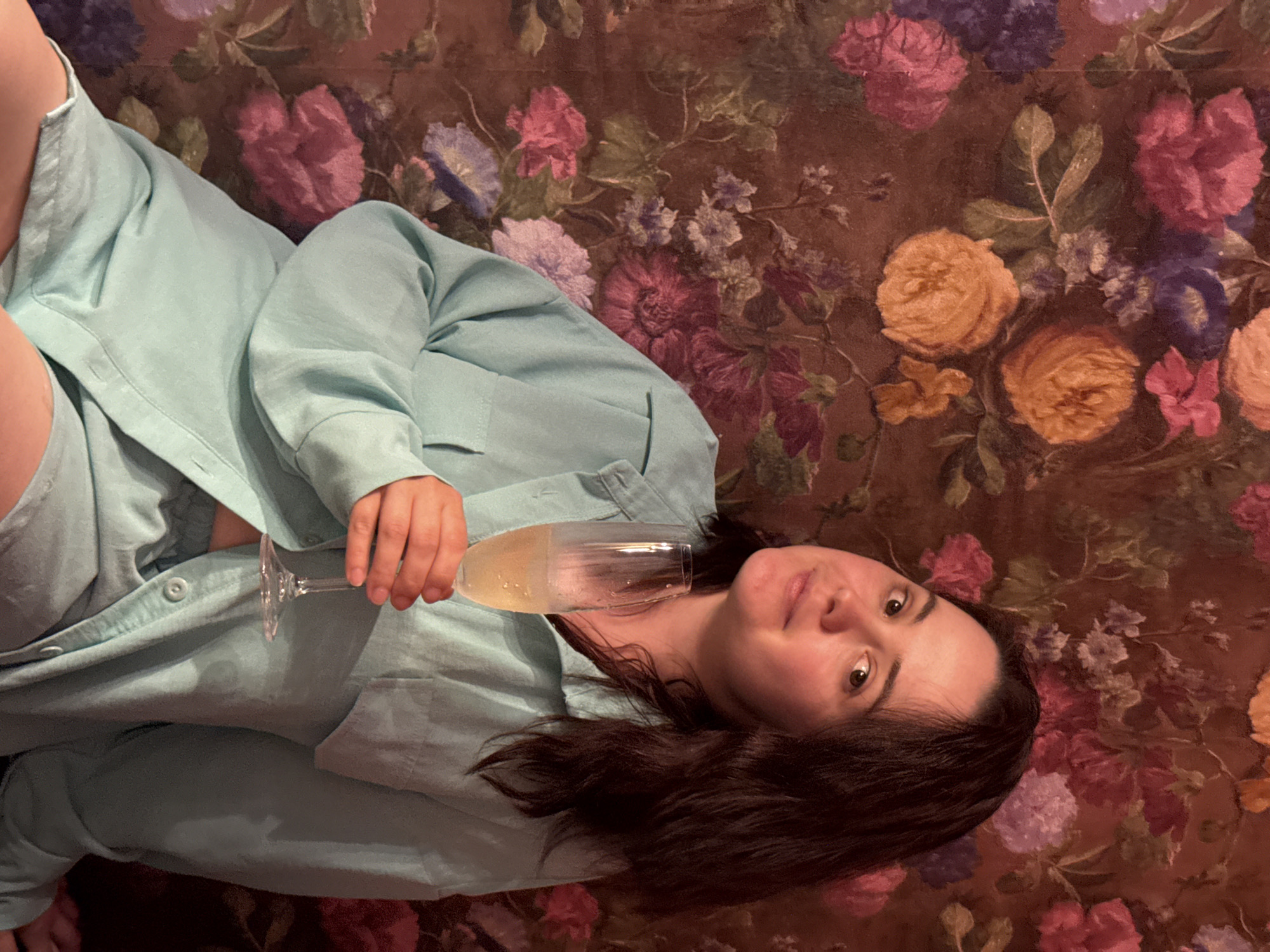 A woman with brown hair is lying down while holding a glass of champagne. She is wearing a light blue pajama set. The background is a wall with a floral pattern featuring roses and other flowers in shades of pink, purple, and yellow. The scene is indoors and appears to be illuminated by artificial light. There is no evidence indicating that the scene took place in the city of Sochi, Russia.