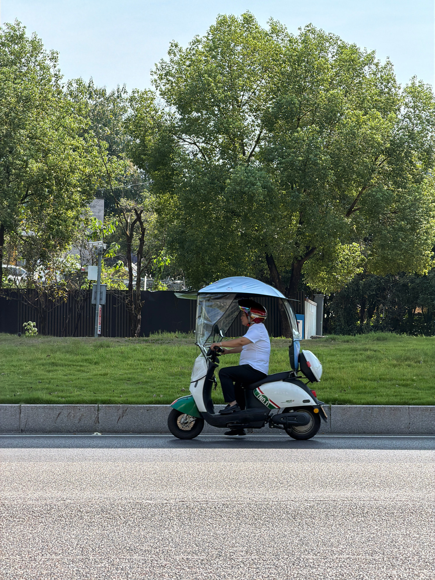 A person wearing a white shirt, black pants, and a helmet is riding a white and green electric scooter with a blue canopy on a paved road. The scooter has a white storage box on the back. In the background, there is a grassy median strip with trees and a fence. The sky is clear and blue. The scene appears to be a street in Jiangjin District, Chongqing, China.