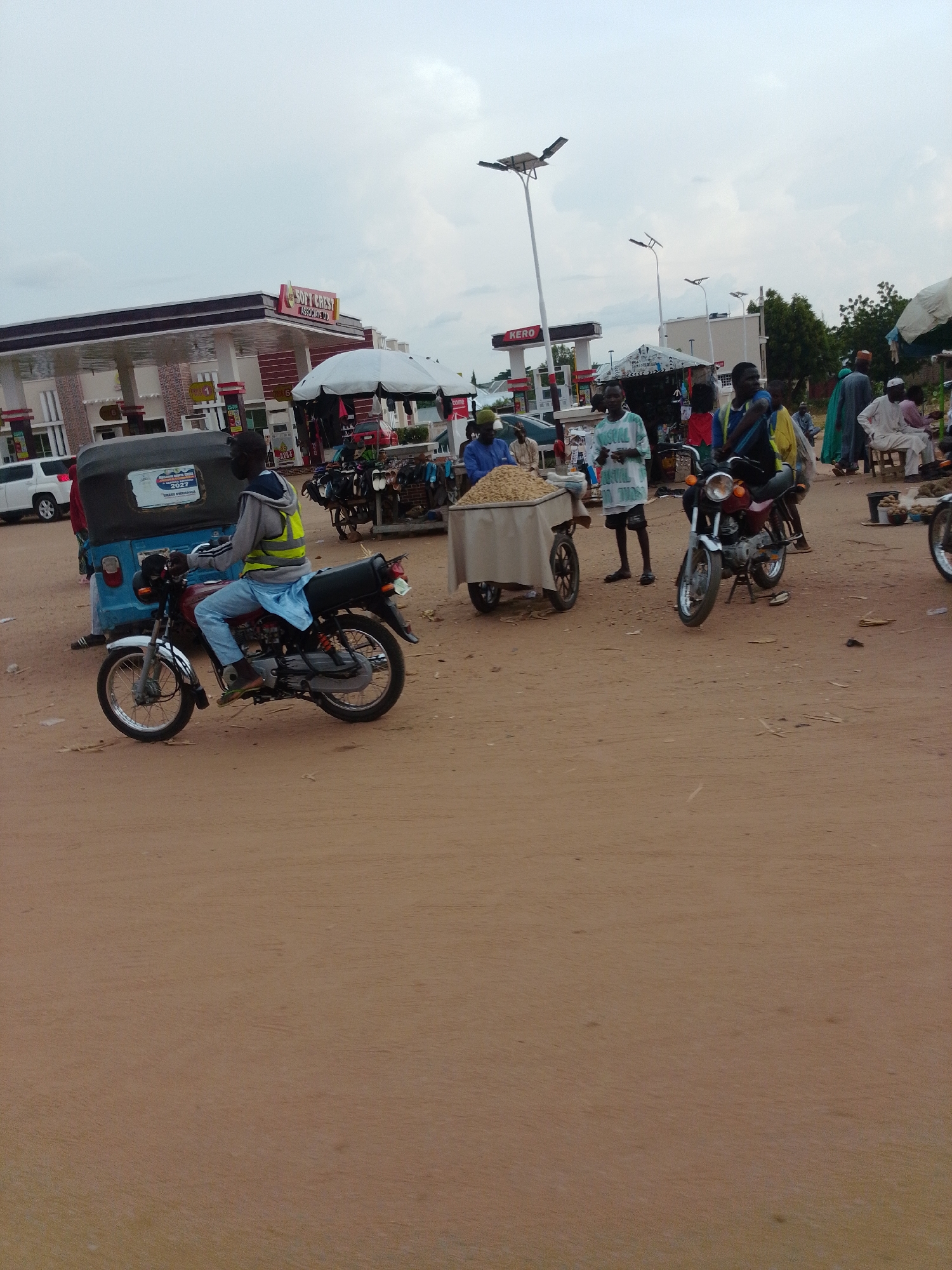 The image depicts a street scene in Gombe, Nigeria, likely a marketplace or a busy thoroughfare adjacent to commercial establishments. In the foreground, a man wearing a reflective yellow vest and light blue trousers rides a maroon motorcycle. He is positioned on the right side of the frame, facing left. Behind him, a blue auto-rickshaw with a black cover is parked.
Further back, a white gas station with a prominent "SOFT CREST ASSOCIATE CO" sign is visible, suggesting a commercial area. Several people are present around stalls set up along the street. One stall features a large cart filled with what appears to be grains or small food items. Other stalls display various goods, including what looks like footwear and clothing. People are seen standing, sitting, and interacting around these stalls.
The environment is characterized by dusty, unpaved ground and a hazy, overcast sky. The presence of the gas station and the organized market stalls suggests an active urban or semi-urban setting. The overall atmosphere appears to be that of a bustling day, with economic activity taking place.
