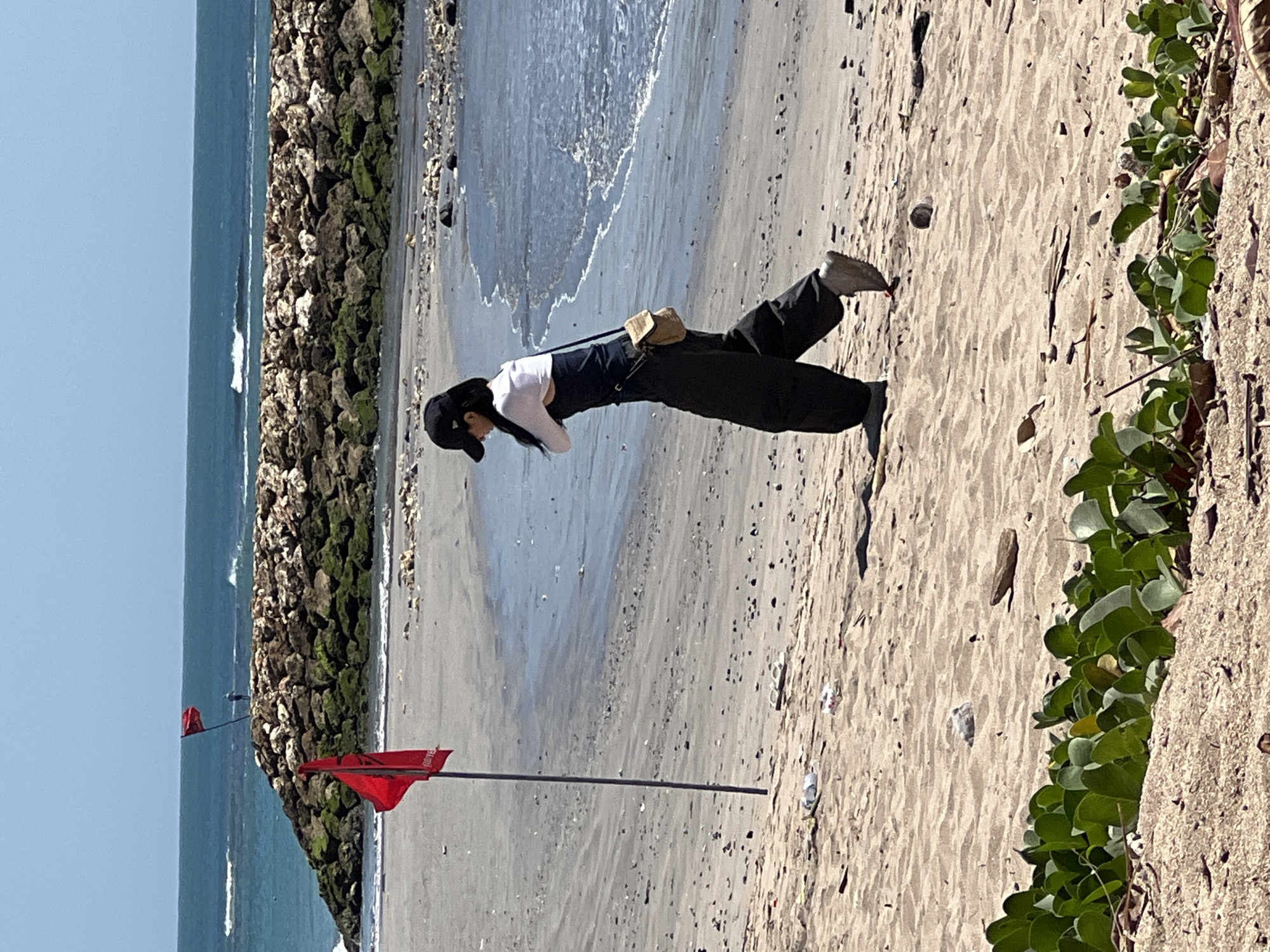 In Kuta, Bali, Indonesia, a woman walks on a sandy beach. The woman wears a black baseball cap, a white shirt, and dark overalls. She carries a small tan bag across her body. In the foreground, green vegetation grows on a sand dune. In the midground, a red flag on a pole stands on the beach. The background shows the ocean with waves breaking against a rocky breakwater. A second red flag is visible on the breakwater. The sky is clear and blue. The beach is littered with debris.
