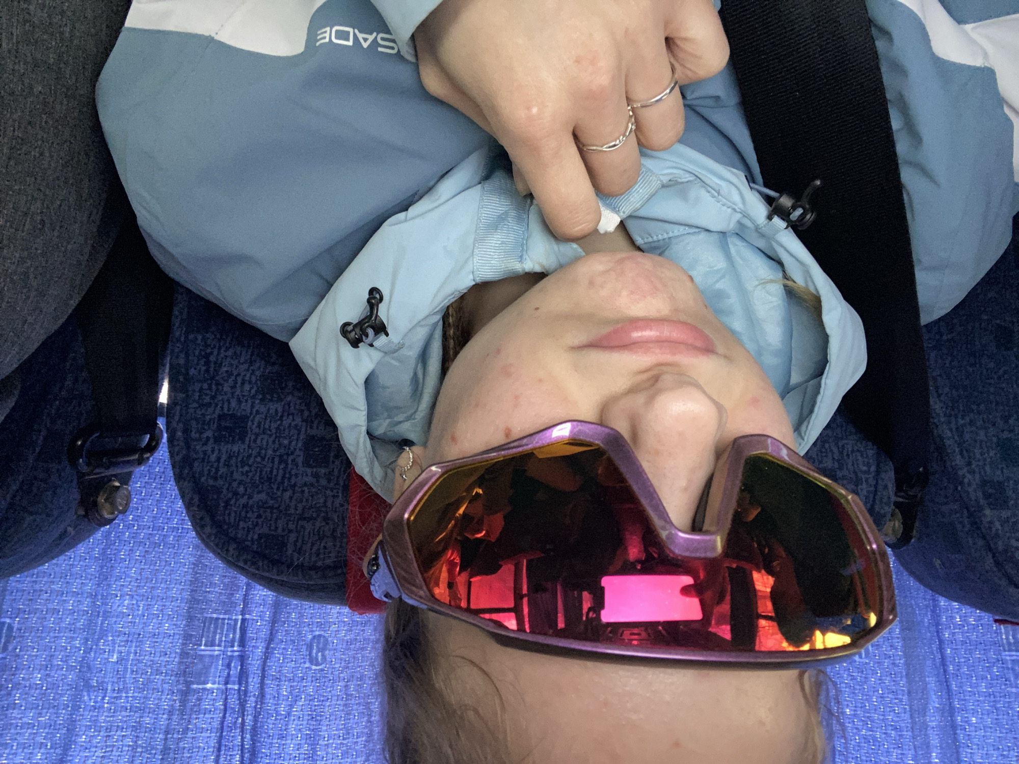 A close-up, inverted photograph shows a person wearing iridescent pink and purple ski goggles. The person is wearing a light blue jacket with a white stripe at the shoulder, and two rings are visible on their hand near their face. The person appears to be seated, as indicated by a seatbelt and upholstered seat in the background. A blue textured surface is also visible. The photograph was most likely taken indoors, potentially on a bus or similar vehicle. The location in the city of Berezniki, Russia is not discernible from the image.