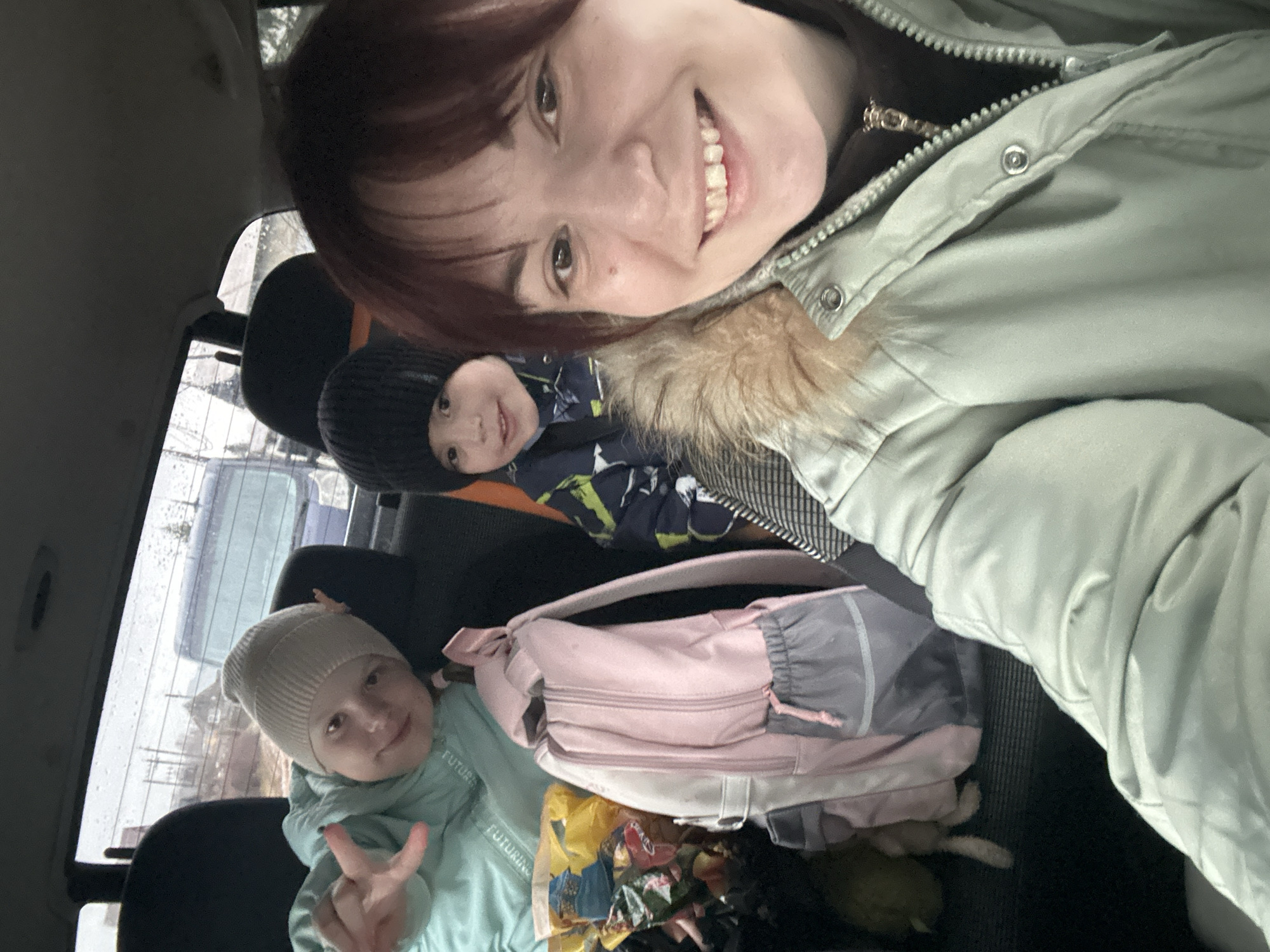 The image shows a woman taking a selfie in what appears to be the back seat of a car, along with two children. One child, wearing a light green jacket and beige hat, is making a peace sign. The other child is behind, wearing a black hat and patterned jacket. A pink backpack and a bag of snacks are in the seat. The window shows a wet, blurred outdoor scene. There is no indication in this image of it being in Фоки, Russia.