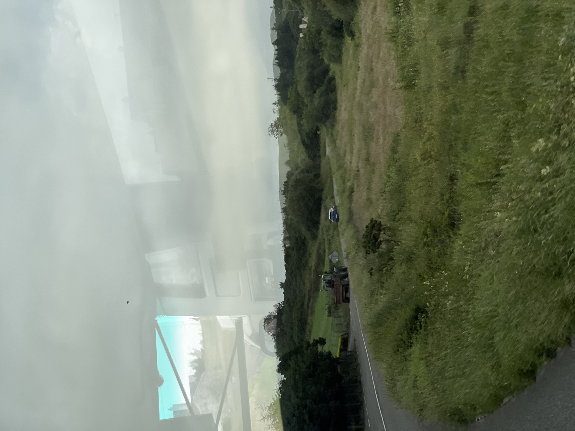 The image shows a rural landscape with a winding road in Falcarragh, Ireland. A green tractor with a trailer is driving on the road, followed by a dark-colored car. The landscape is characterized by rolling hills covered in lush green grass and scattered trees. The sky is overcast with clouds, suggesting a typical Irish day. Reflections of a building and the sky are visible on the surface through which the photo was taken, likely a window.