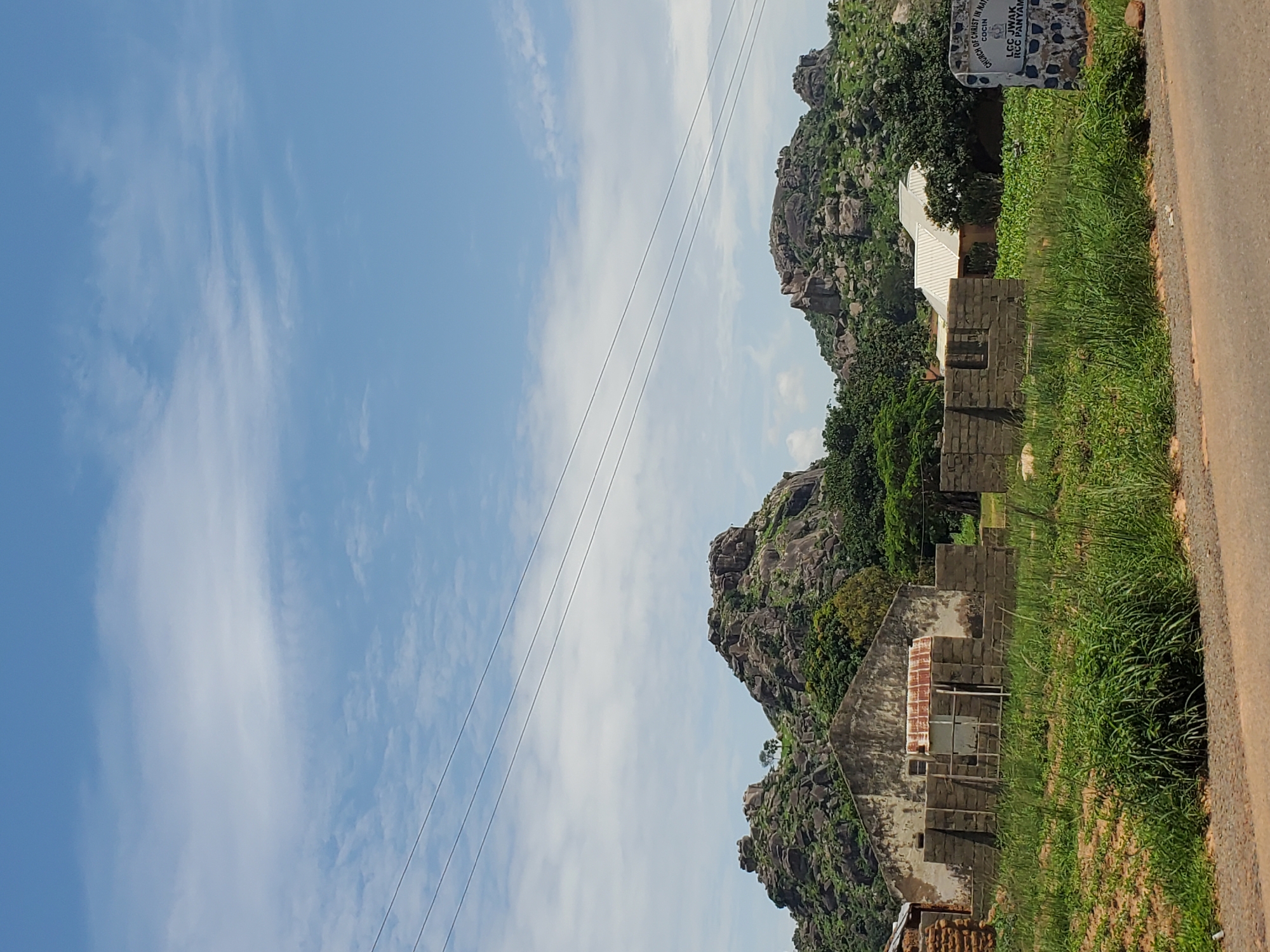 The image depicts a rural scene in Panyam, Nigeria, characterized by rocky hills in the background. In the foreground, there is an unfinished brick structure, suggesting ongoing construction or a derelict building. A partially visible house with a rusty corrugated iron roof stands to the left of the unfinished structure. To the right, a white roofed building is partially obscured by vegetation. A stone monument or signpost is also visible on the right side of the image, with text that appears to read "CHURCH OF CHRIST IN NIGERIA COCHIN LCC Jwak RCC Panyam." The environment is overgrown with grass and foliage, and power lines stretch across the sky. The sky is partly cloudy.