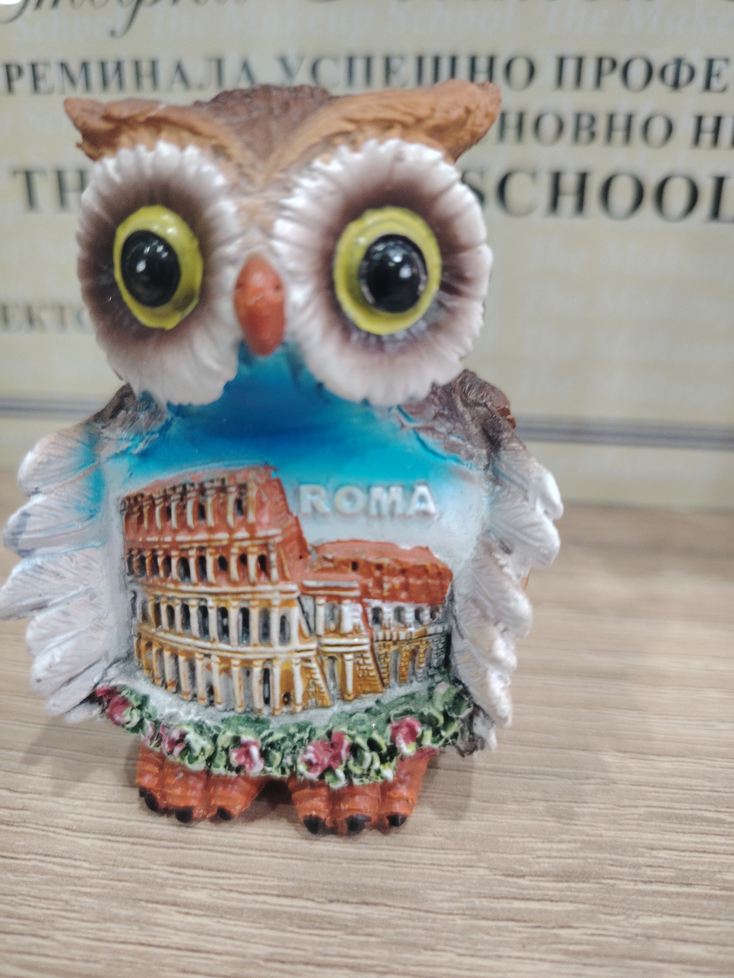 A small, decorative owl figurine is the central subject of this image. The owl is made of a material that appears to be ceramic or resin and is designed to resemble a real owl with its large, round, yellow eyes with black pupils, a pointed beak, and feathery details.
The owl is decorated with a scene depicting the Colosseum in Rome, Italy. The architectural structure of the Colosseum is visible in vibrant detail, colored in shades of red and brown. Above the Colosseum, a blue sky is depicted. The word "ROMA" is written in white capital letters across the blue sky portion of the decoration. The base of the figurine is adorned with a band of green foliage and small red flowers, and the owl's feet, which are orange with black claws, are visible at the bottom.
The figurine is placed on a light brown wooden surface, likely a table or shelf. In the background, there is a blurred image of text printed on a light-colored surface. Some of the visible text in the background appears to be in Cyrillic script, with letters like "РЕМИНАЛА," "УСПЕШНО," "ПРОФЕ," "НОВНО," and "SCHOOL." The overall lighting suggests an indoor setting, possibly during the daytime. The image is a close-up shot, focusing entirely on the owl figurine and its immediate surroundings. There are no people or animals present in the scene, and no specific activity is taking place other than the display of the object. The context of Haskovo, Bulgaria, is provided, but there are no visual cues within the image itself that specifically point to this location.