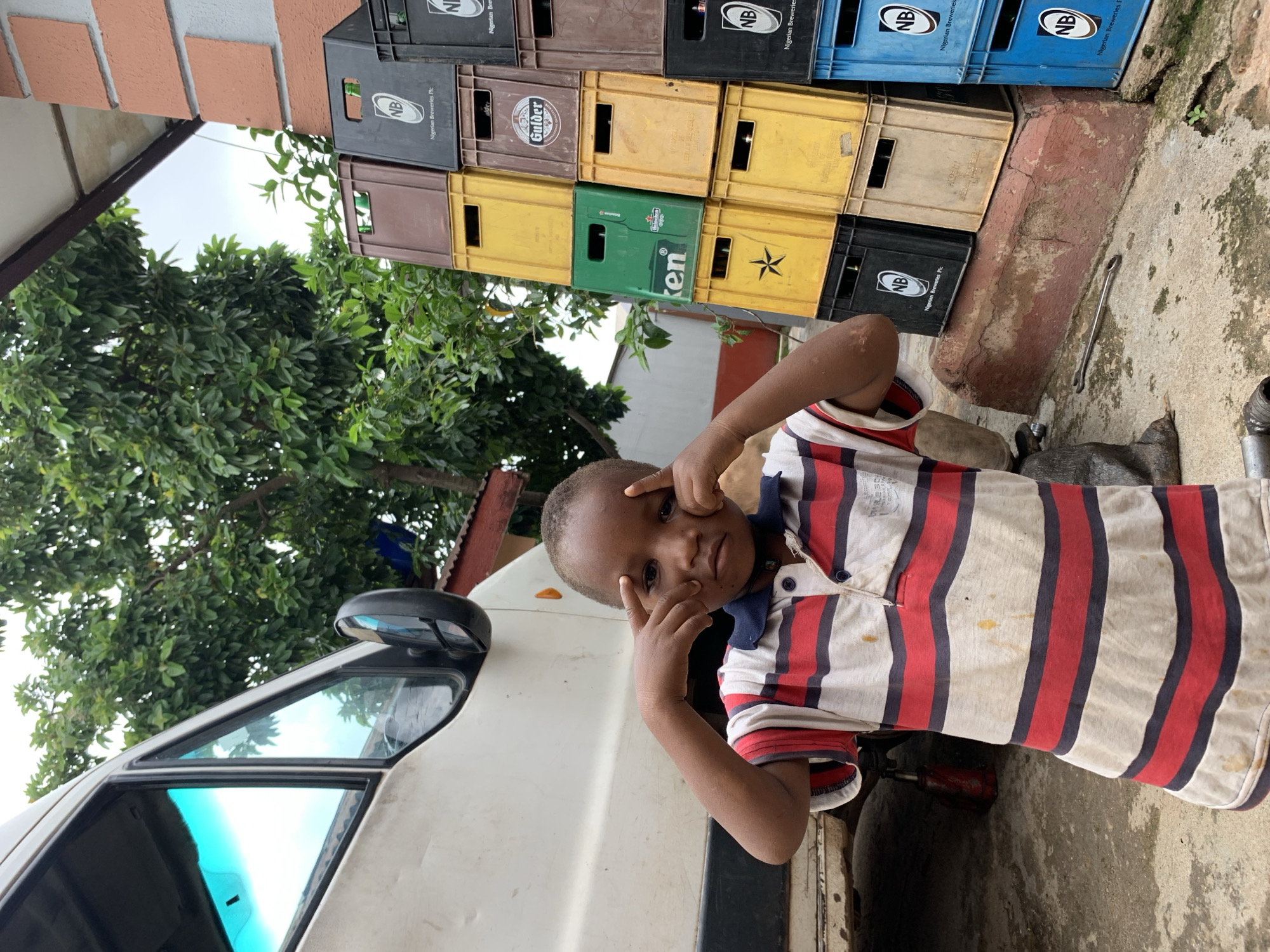 This is a portrait-oriented, outdoor photograph taken in Jos, Nigeria. The primary subject is a young boy, likely of African descent, with dark skin and short hair. He is positioned in the foreground, leaning against a white vehicle, and looking directly at the camera with a playful or perhaps slightly mischievous expression, making a "peace" sign with his fingers near his face. He is wearing a striped polo shirt with red, navy blue, and cream-colored bands. Stains are visible on the shirt.
Behind the boy and the vehicle, a stack of plastic crates is visible. These crates are colorful, with some being yellow, brown, black, and blue. Some of the crates have logos and text on them, including "NB" and "Nigerian Breweries Pic," as well as "Gulder" and what appears to be a Heineken logo on a green crate. The crates are stacked outdoors, suggesting a storage area or a marketplace.
To the left of the boy and the crates, there is lush green foliage from a tree, and a portion of a building with reddish-brown bricks and a white overhang is visible at the top left of the frame. The ground beneath the crates and extending to the right is a concrete or stone surface, with some visible cracks and a small plant growing in one of them. A metal wrench is lying on the ground near the crates.
The lighting suggests it could be daytime, possibly overcast, given the diffused light and the brightness of the sky. The overall scene depicts a candid moment with a child in a somewhat utilitarian or industrial setting.