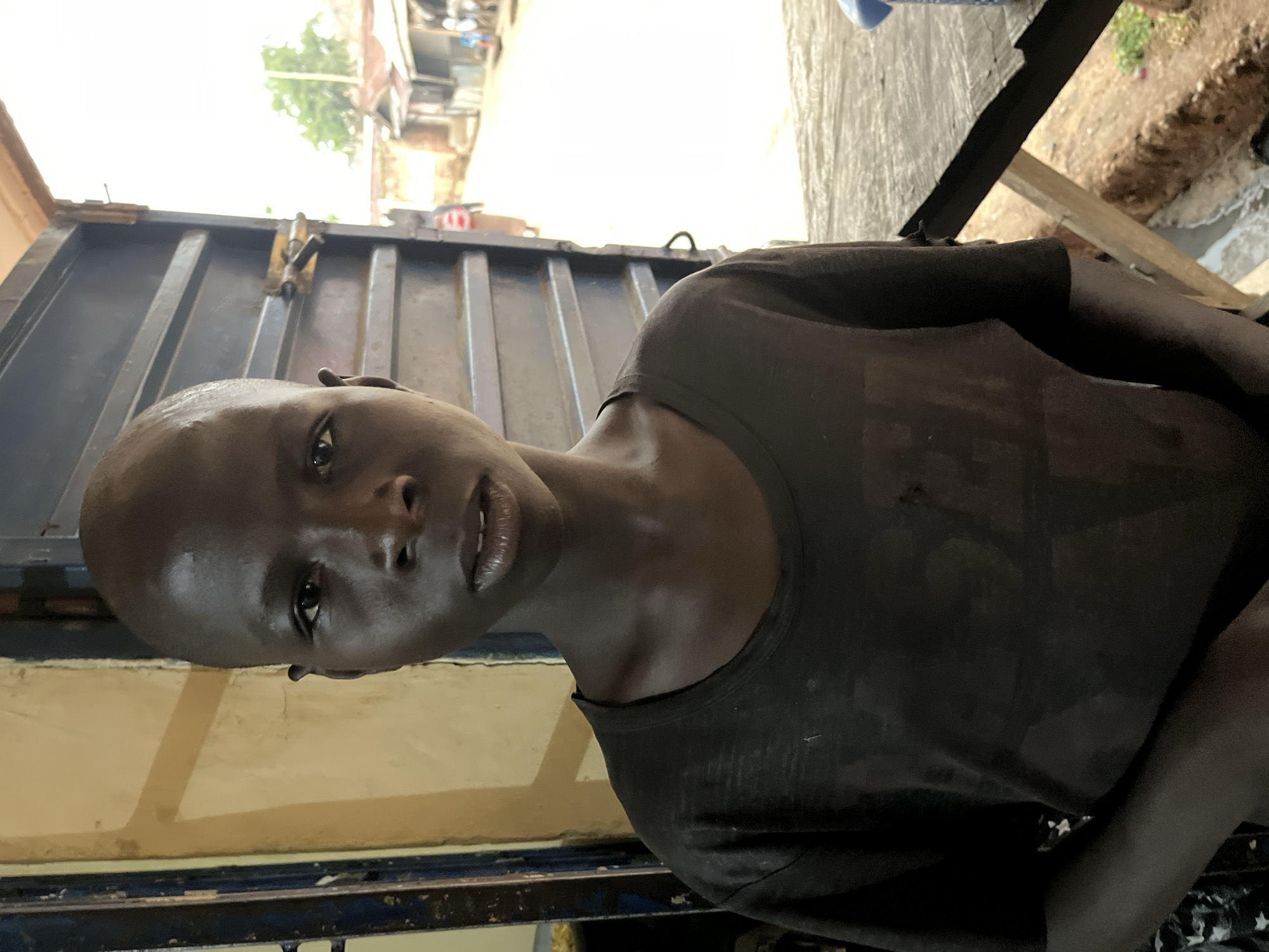 The image shows a close-up of a young, dark-skinned person wearing a black t-shirt. The person is partially indoors, standing next to a corrugated metal wall painted with yellow and blue trim. The background is blurry but appears to show a street scene, suggesting a location in the city of Jos, Nigeria. Visible in the background are building exteriors, a tree, and other unidentifiable objects. The person is looking directly at the camera.