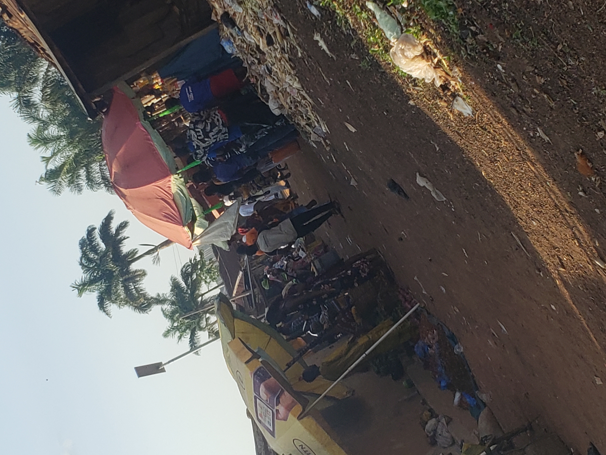The image depicts an outdoor commercial area or market street in Gumel, Nigeria, under a bright, clear sky. In the upper background, several prominent palm trees are visible. A tall pole with a rectangular panel, possibly a solar light or sign, stands in the left mid-ground.
The scene is populated by numerous individuals, mostly adults, engaged in various activities typical of a market. Two large market umbrellas dominate the mid-ground: one is red with a green valance, positioned centrally to the right, beneath which several people are gathered. The other, on the left, is yellow with "MTN" branding and advertisements, sheltering additional individuals.
People are dressed in casual attire, including t-shirts, patterned shirts, and robes. Actions include standing, walking, and interacting near the market stalls. One person in a yellow garment is partially visible beneath the MTN umbrella, facing left. Another individual in a blue shirt is prominent near the red umbrella.
The ground is unpaved, consisting of reddish-brown dirt or dry mud, showing signs of heavy foot traffic. Extensive litter and debris are scattered across the ground, particularly in the right foreground and along the edge of the street, including numerous plastic bags, paper, and other refuse. A distinct shadow line diagonally crosses the foreground from left to right, indicating direct sunlight from the upper right. A rustic wooden or corrugated metal structure forms the right boundary of the scene, while a more solid wall-like structure is on the left.