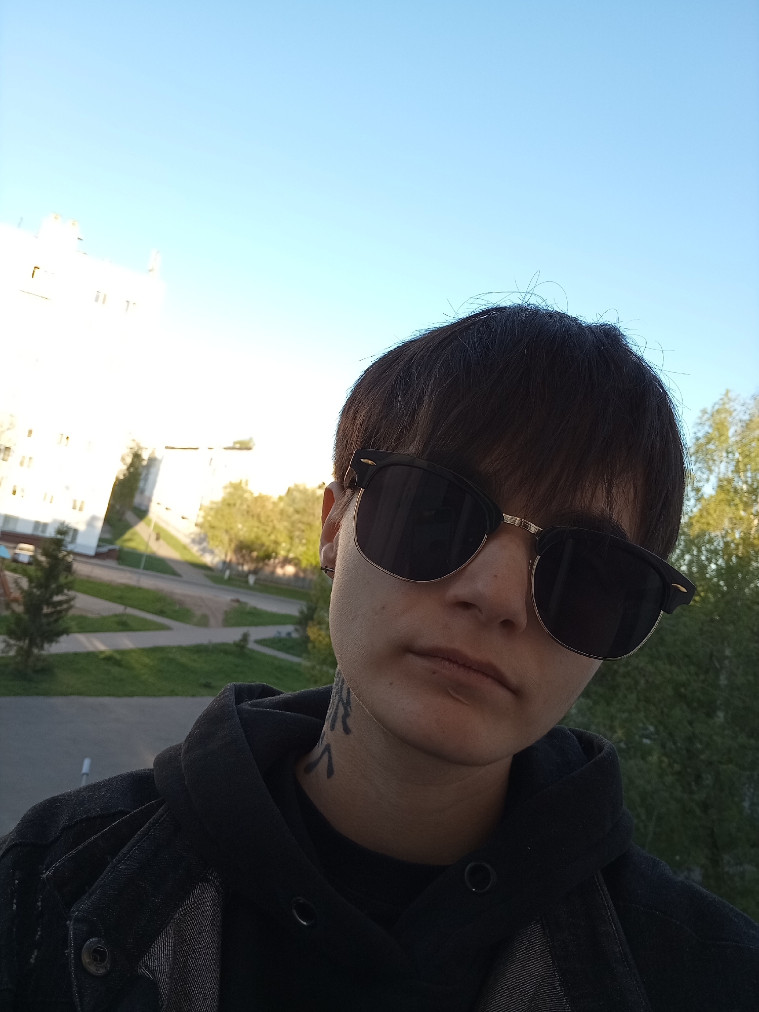 A selfie of a person with dark brown hair, wearing sunglasses and a black hooded jacket, with a tattoo on their neck. They are outdoors, likely on a balcony or elevated position. The background shows an urban environment with a multi-story white building, green trees, and what appears to be a sidewalk or paved area. The sky is clear and blue. Based on the style of architecture and general appearance, the location is consistent with urban areas in Naberezhnye Chelny, Russia.