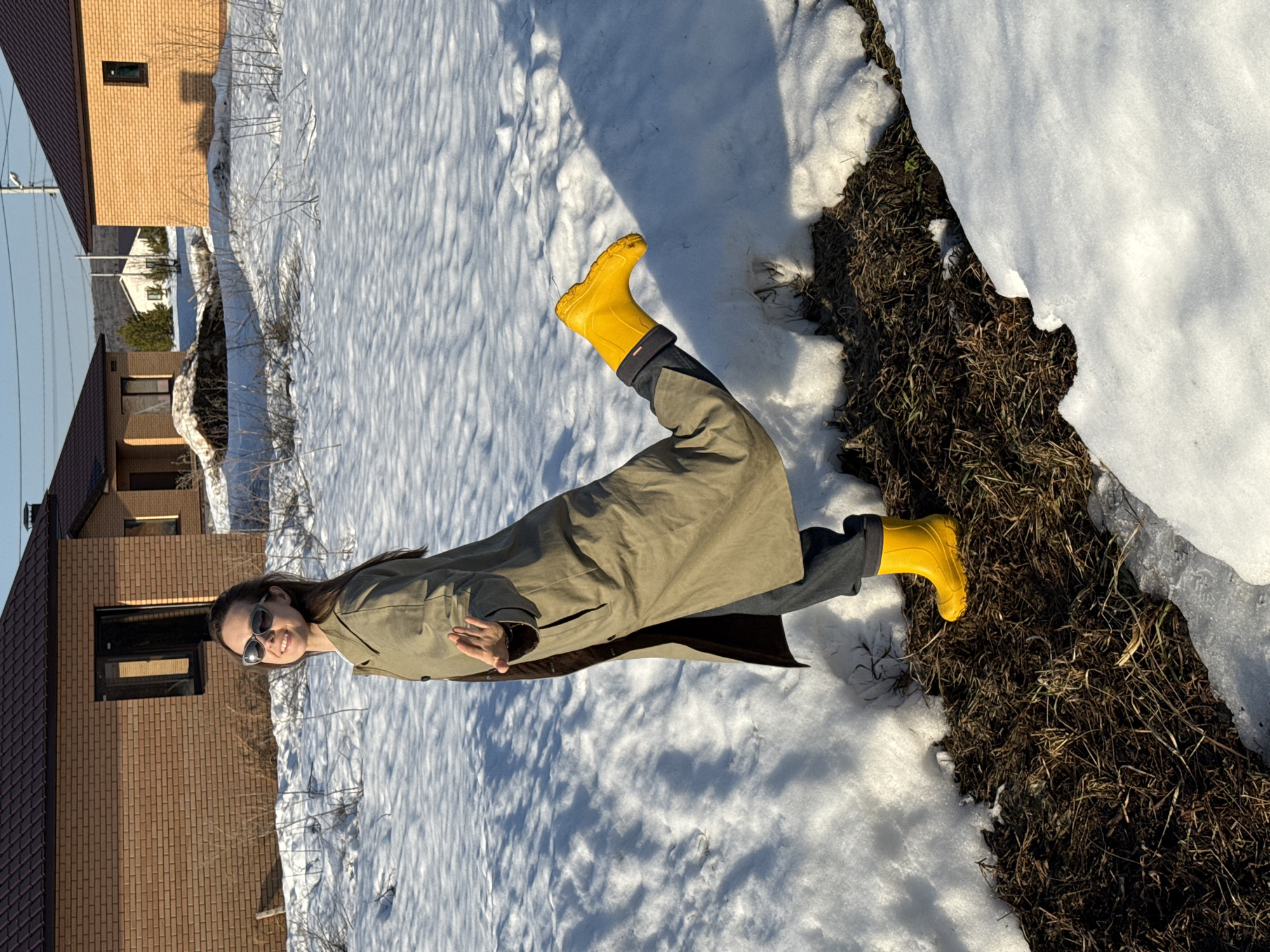 An outdoor scene in Chaikovsky, Russia, depicts a female subject posing in a patchy snow-covered field under bright sunlight. The woman, appearing to be an adult, stands centrally, turned slightly to her right, looking directly at the camera with a smile. She has long brown hair and wears dark sunglasses, an olive-green long trench coat, dark pants, and bright yellow rain boots. Her left leg is bent at the knee, raising her left foot backward, while her right arm is extended forward with an open palm.
The immediate foreground consists of a strip of exposed dark brown soil and dried vegetation, bordered by irregular patches of white snow. The ground beyond the woman is predominantly covered in snow, showing texture from footprints or melt patterns, with scattered patches of dry, brown vegetation.
In the background, two brick-clad residential structures with dark brown roofs are visible. The house to the left features a dark-framed window. The house to the right has a similar dark-framed window and casts a shadow on the snow. A third, smaller, white building is discernible between these two structures, further in the distance. Power lines and utility poles are present in the sky above the houses. A distant, possibly forested, hill or mountain range is visible on the horizon. The environment indicates a late winter or early spring setting with melting snow.