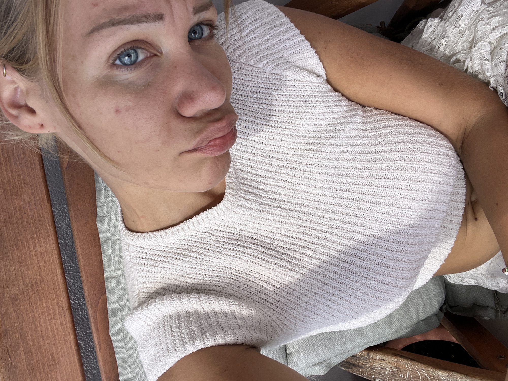 A young Caucasian woman with blonde hair and blue eyes is captured in a close-up, overhead shot. She is wearing a white knitted top and is seated on a chair with a light green cushion. The background consists of wooden planks. The lighting suggests an outdoor setting, possibly a patio or garden, bathed in natural sunlight. The woman's expression is pensive, with a slight pout. The image is taken from a high angle, looking down at the woman. There is no discernible city or specific location information present in the image itself.